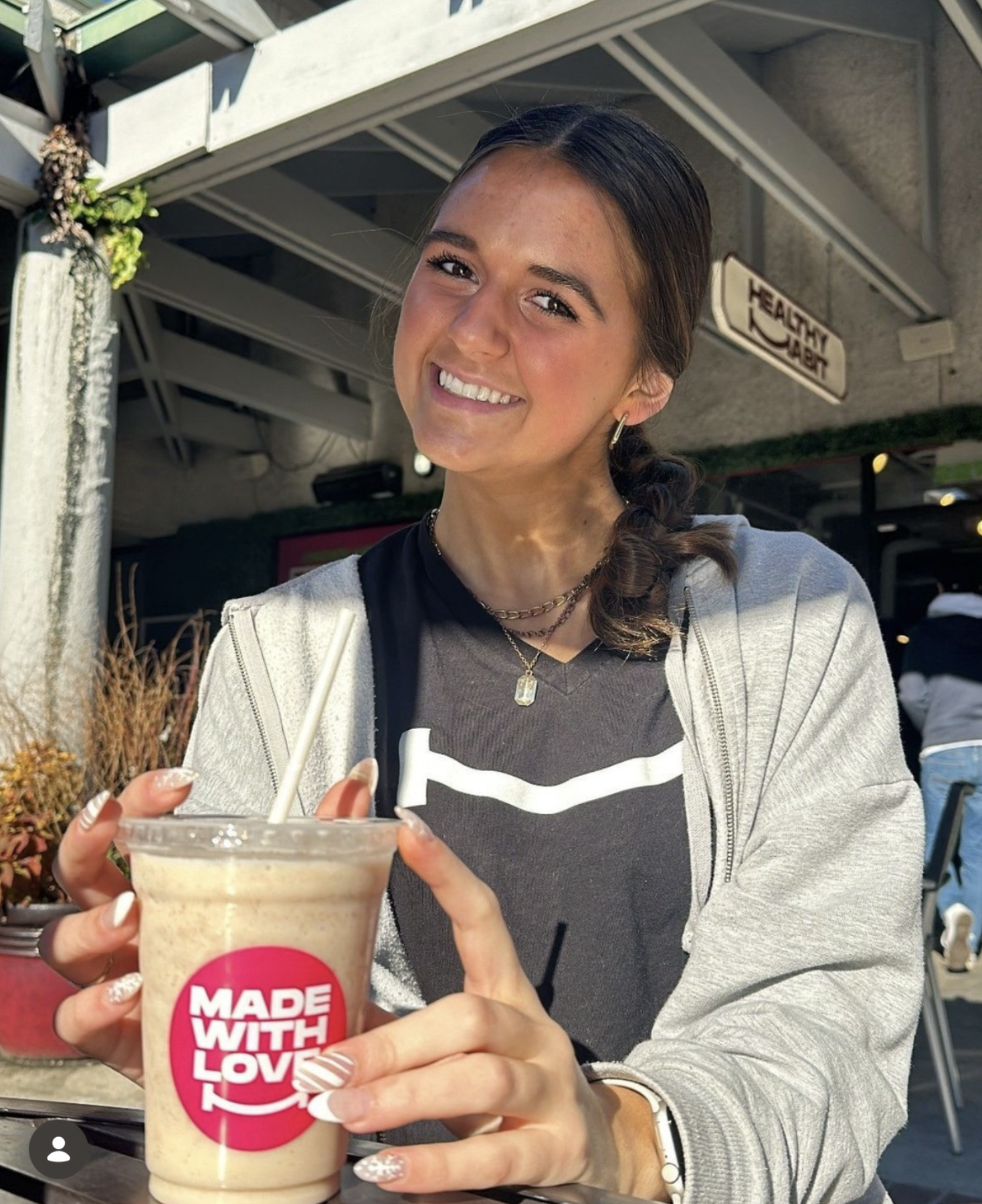 A young woman smiling while sitting outdoors, holding a blended coffee drink in a clear cup with a pink sticker that says "Made with Love". She has dark hair tied back, wears a black t-shirt with white stripes, a gray zip-up hoodie, layered necklaces