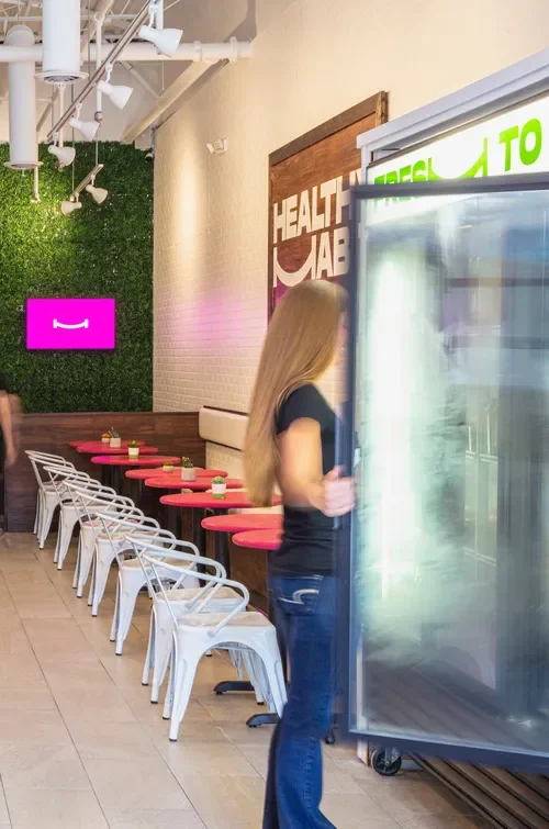 A woman with long blonde hair wearing a black shirt and blue jeans stands near a glass refrigerated display case in a cafe or restaurant. The interior features pink tables with white chairs, a green wall with white lighting fixtures, and a sign that 