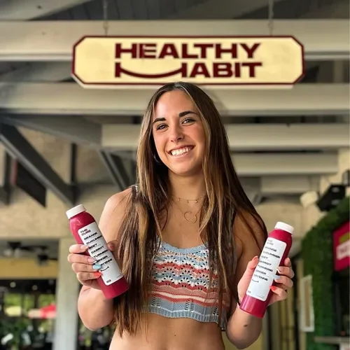 Smiling young woman holding two pink bottles of health supplements in front of a sign that says 'Healthy Habit'.