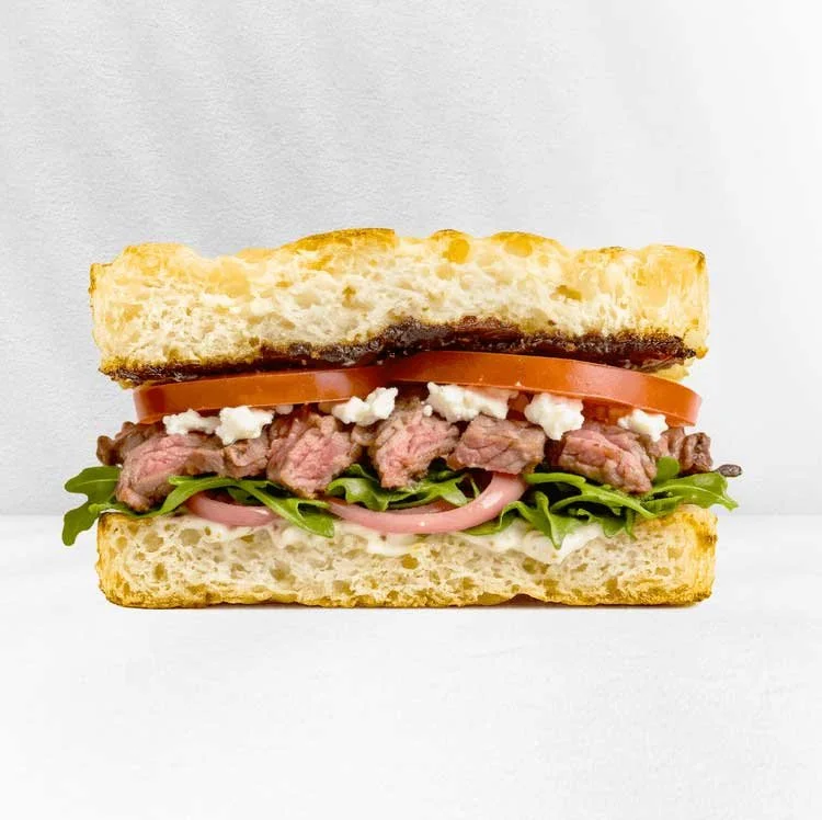 A sandwich with sandwich bread, tomato, beef steak, feta cheese, lettuce, and onions.