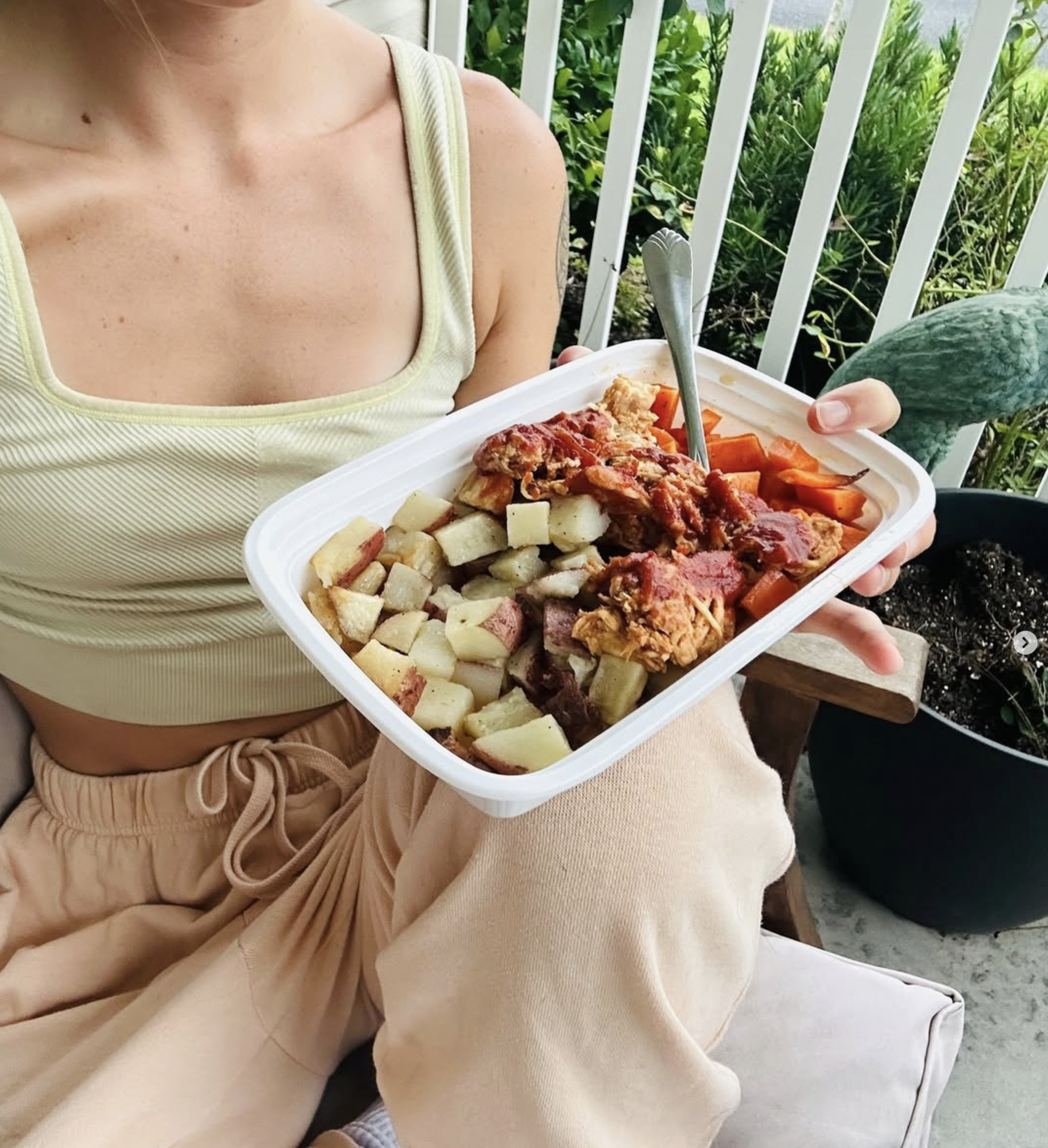 Person in a yellow tank top holding a container of food with baked potatoes, vegetables, and meat, sitting on a porch with greenery in the background.