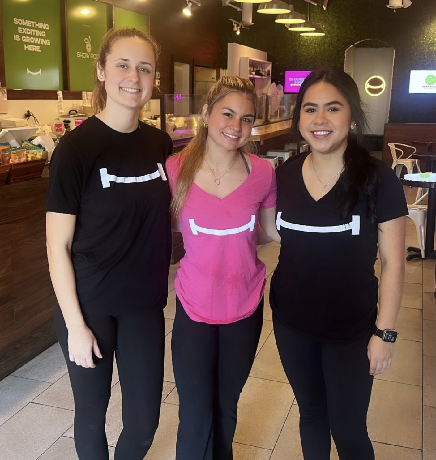 Three women standing together in a restaurant or cafe, smiling at the camera. They are wearing black and pink T-shirts with a white bracket symbol on the front.