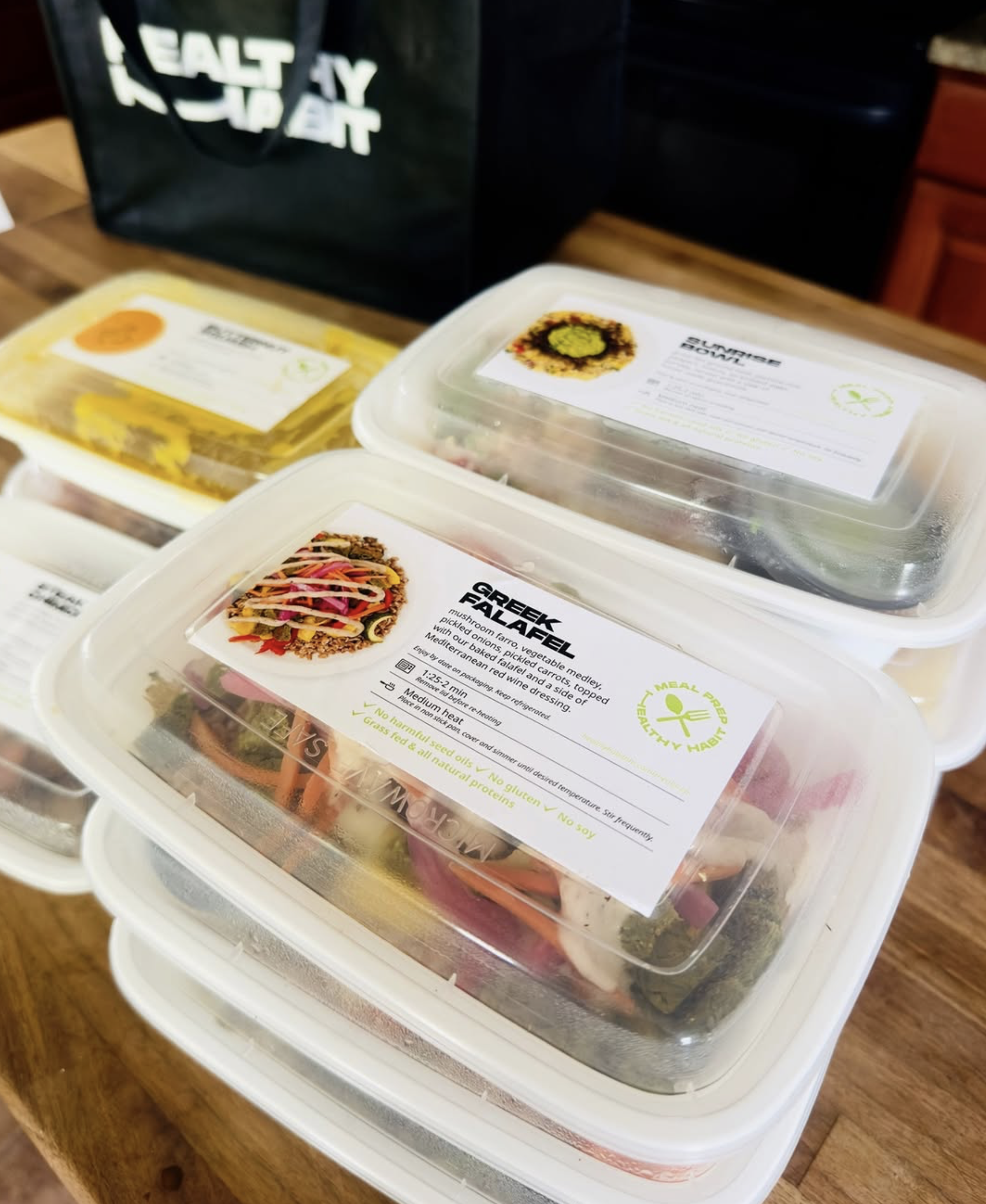 Three plastic food containers on a wooden table, each with a label showing different meal options: one with a colorful Greek falafel salad, another with a vegetable medley, and the third with a dish containing noodles and vegetables. There is a black