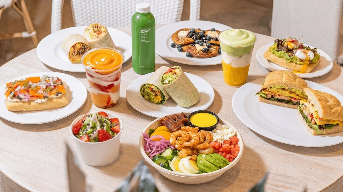 A table with various dishes including wraps, salads, smoothie drinks, pastries, and desserts at a restaurant.
