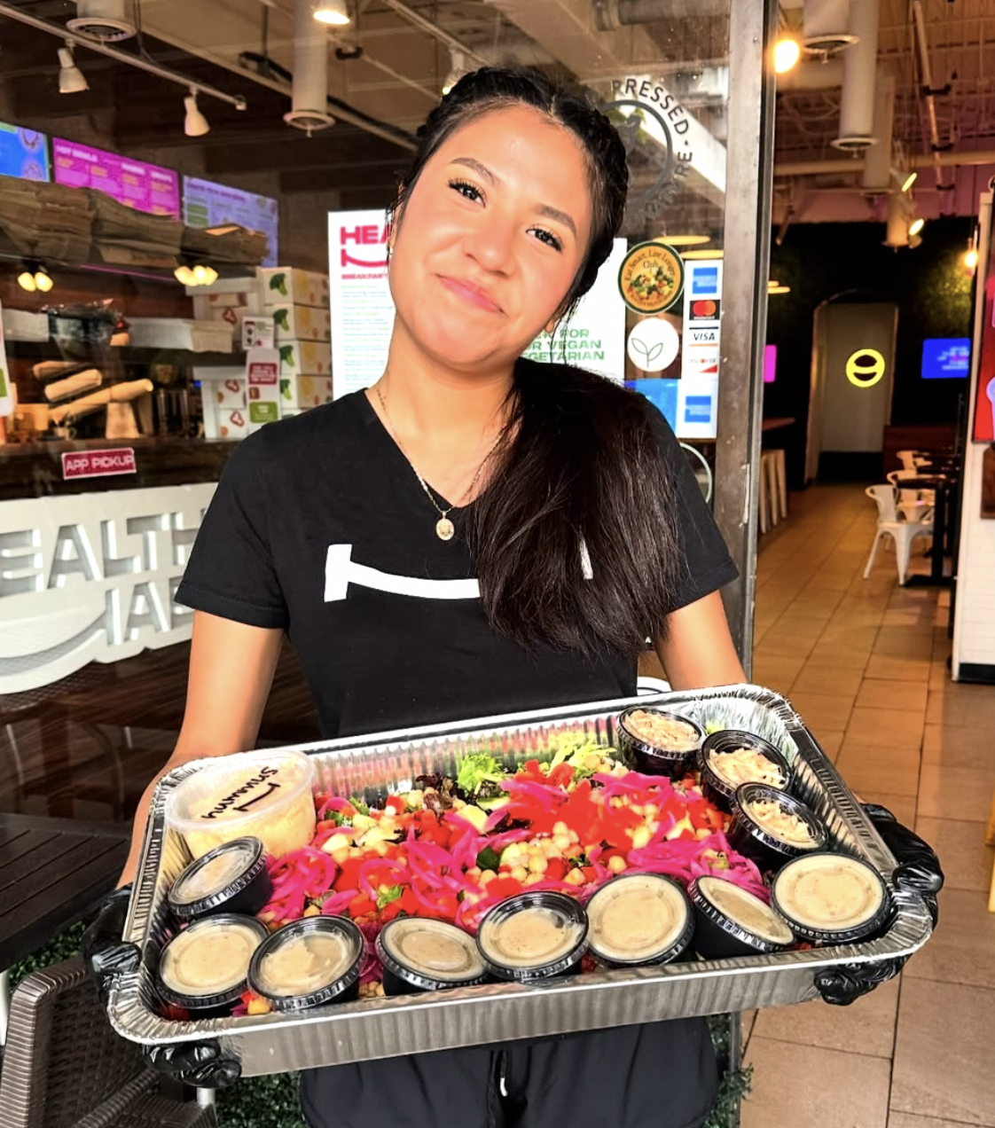 A woman smiling and holding a tray of colorful salad with edible flowers and small containers of dressing inside a restaurant.
