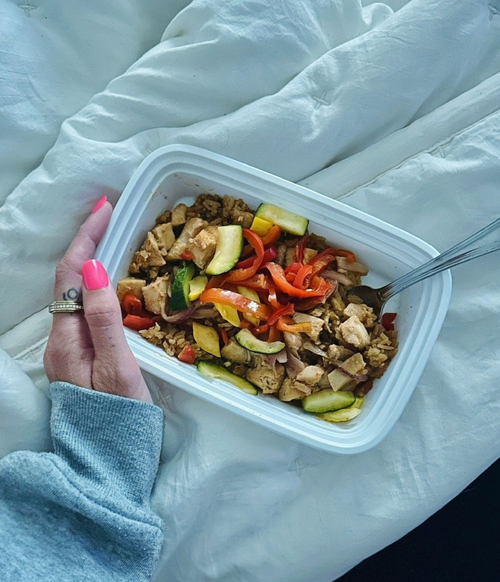 Hand holding a container of stir-fried vegetables and tofu on a bed with white sheets