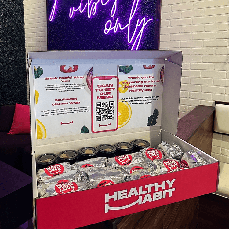 A red and white box labeled 'Healthy Habit' contains wrapped food items with 'Made with Love' stickers, kitchen sauce jars, and a QR code for menu ordering. The box is on a wooden table against a brick wall, with a neon sign above reading 'Vibes Only