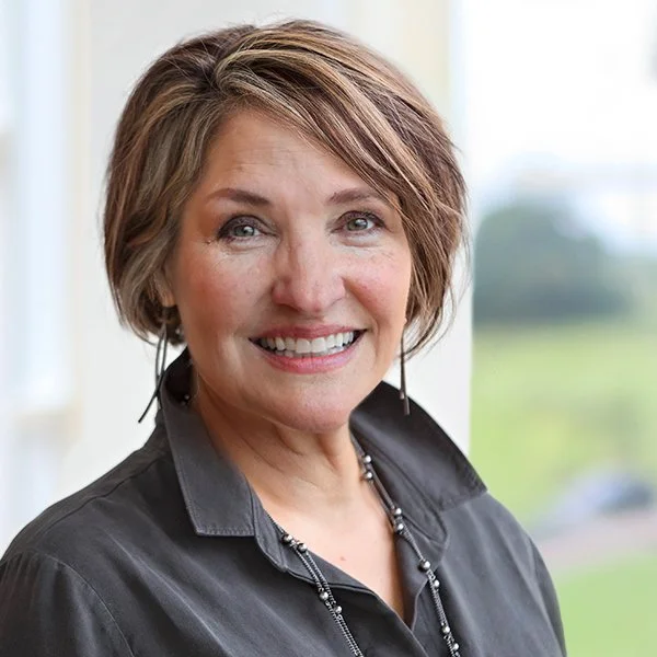 A middle-aged woman with short brown hair and blue eyes smiling, wearing a dark button-up shirt and a necklace, standing indoors near a window with a blurred outdoor landscape in the background.