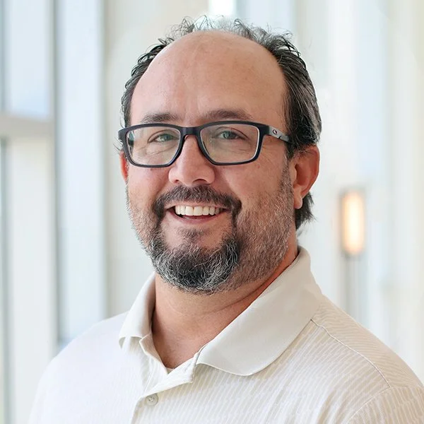 A smiling middle-aged man with glasses, a beard, and mustache, wearing a white polo shirt, standing in a bright indoor setting with windows in the background.