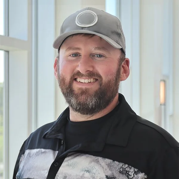 A man with a beard smiling, wearing a gray baseball cap and a black jacket, standing in front of large windows.
