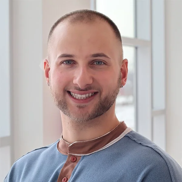 A smiling man with short hair and a beard, wearing a blue shirt with a brown collar, standing indoors near large windows.