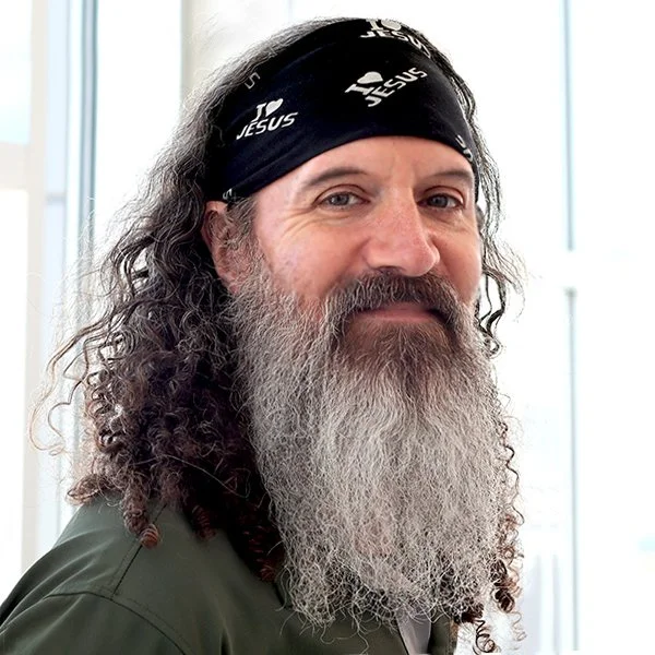 Man with long curly hair and a gray beard wearing a black headband with 'Jesus' written on it.