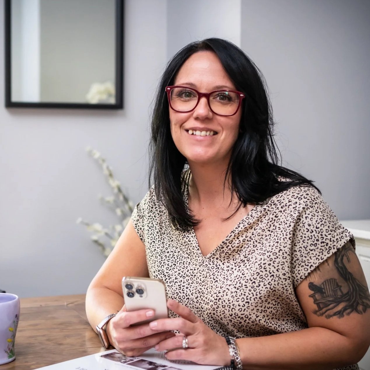 A woman with black hair, glasses, and a tattoo on her arm sitting at a wooden table holding a smartphone, smiling at the camera inside a room with a window, mirror, and decorative items.