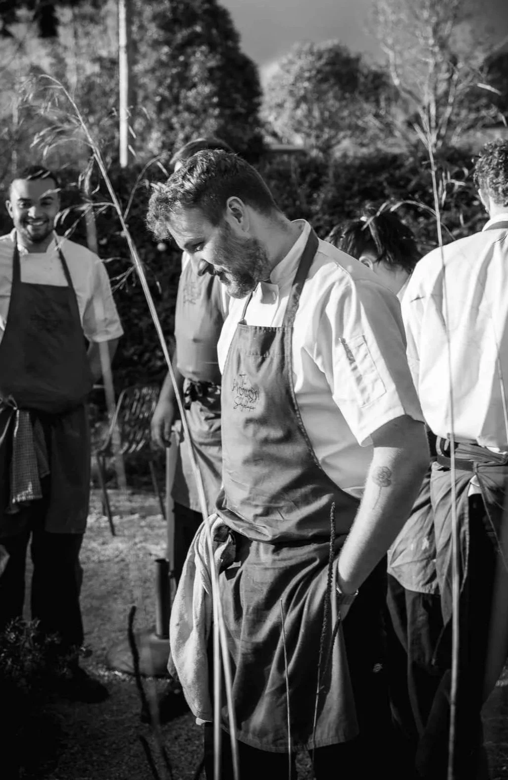 Black and white photo of male chefs wearing aprons and chef jackets, standing outdoors among trees, with one chef in the center looking down and smiling.