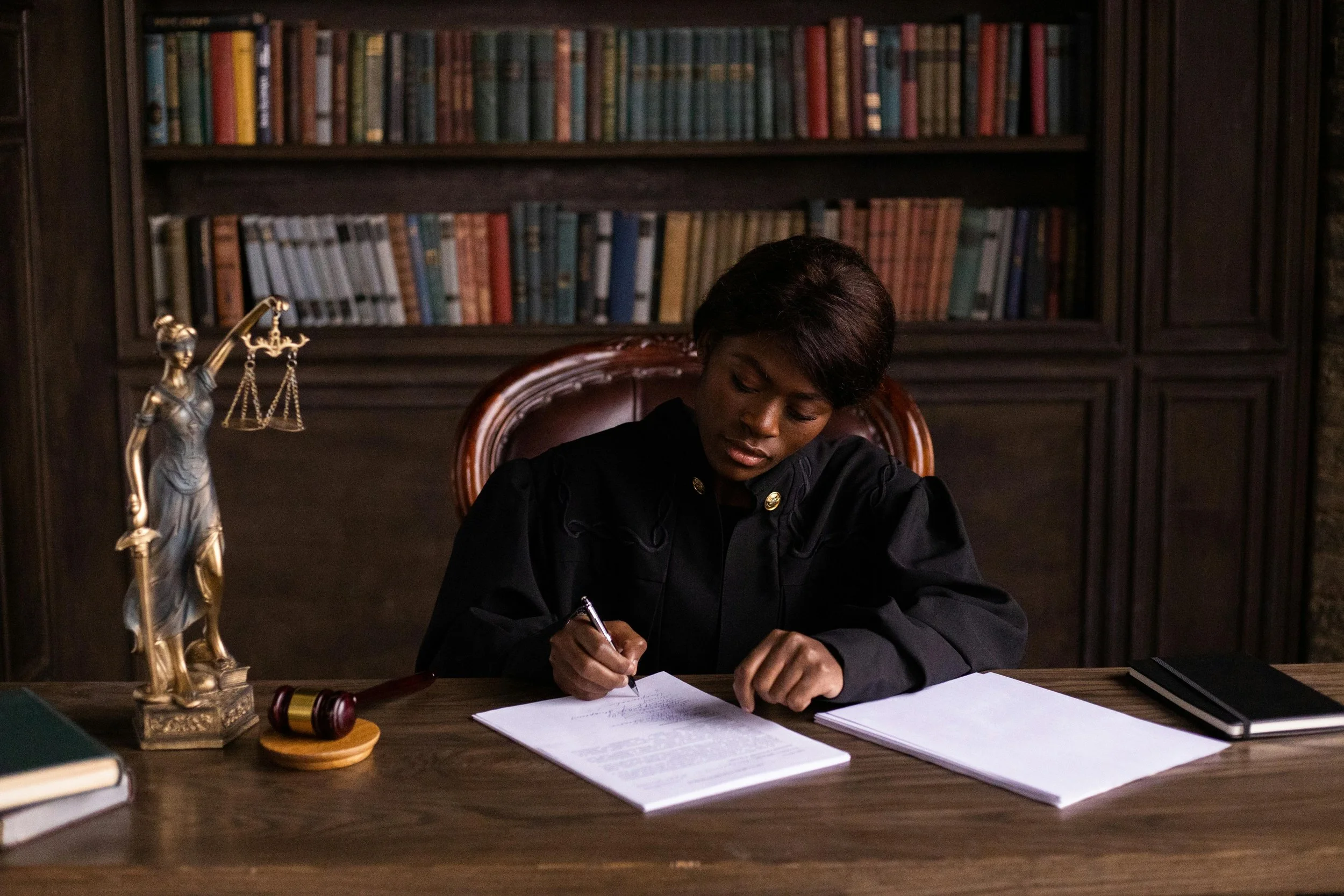 Woman in black judge's robe sitting at a wooden desk with legal papers, gavel, and scales of justice sculpture, in front of a bookshelf filled with books.