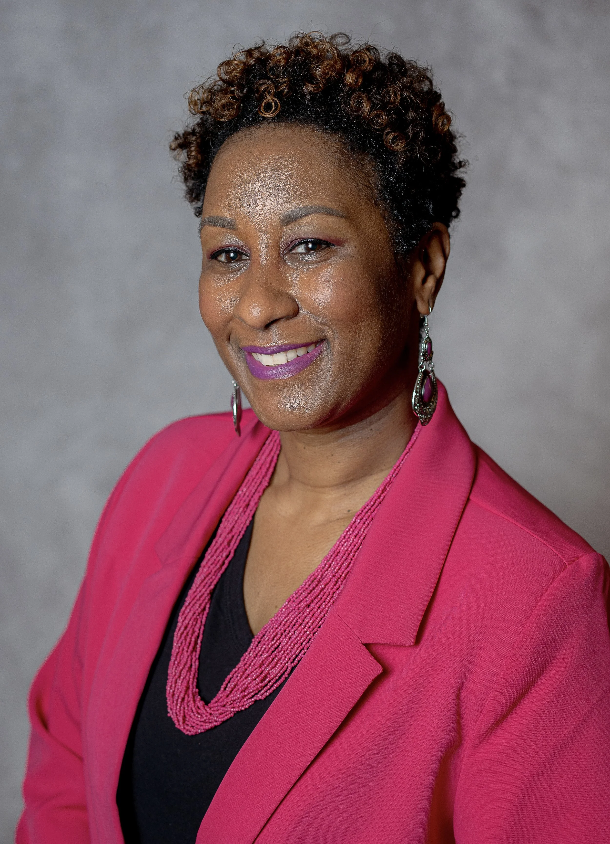 A woman with short curly hair wearing a pink blazer, black top, pink beaded necklace, and matching earrings, smiling at the camera against a gray background.