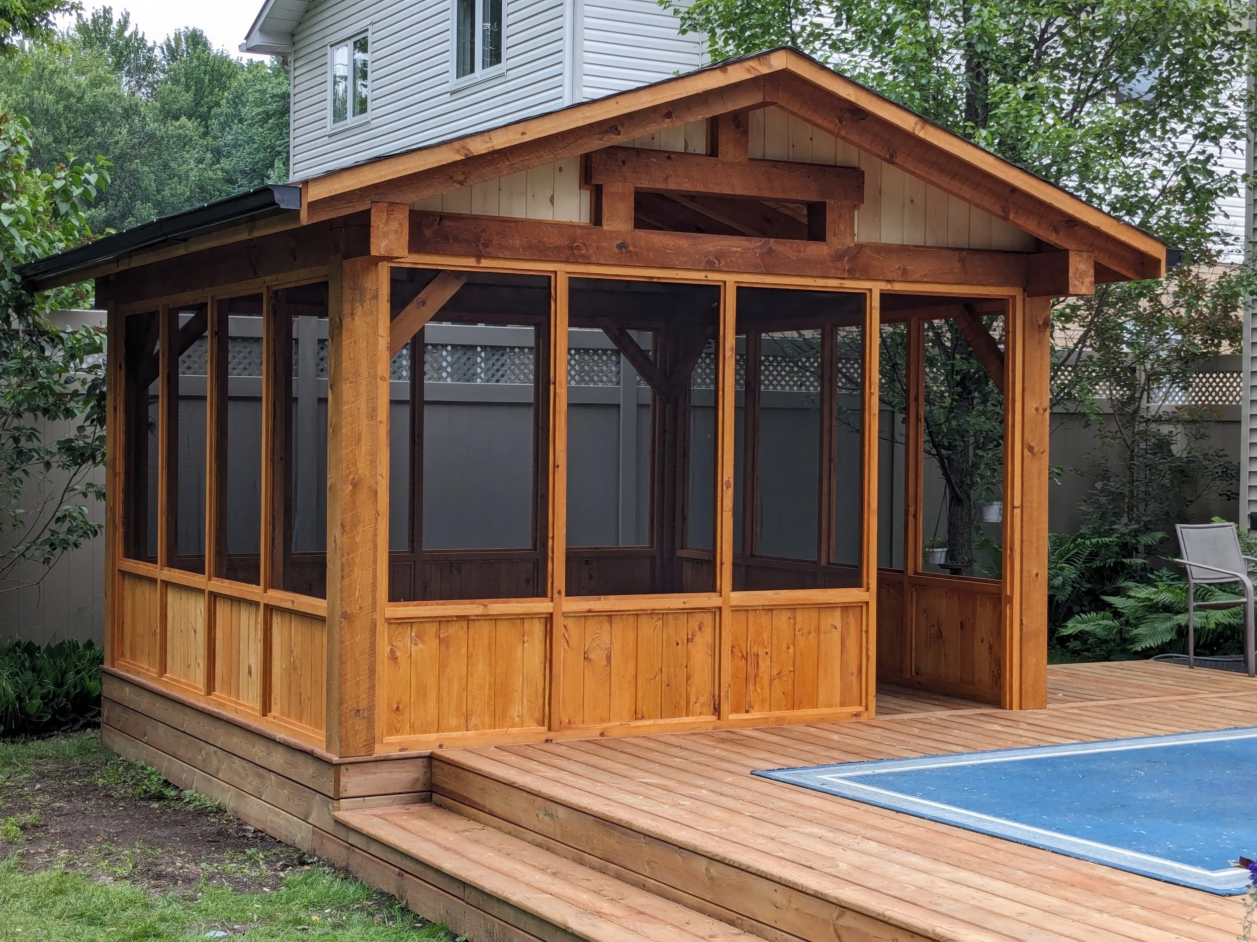Wooden gazebo with screened walls on a wooden deck in a backyard, surrounded by trees and a fence.