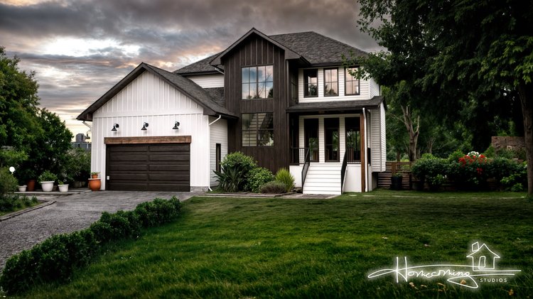 This image features a modern farmhouse-style home with a stunning mix of materials and contrasting tones. The house combines white board-and-batten siding with dark-stained wood accents, creating a dramatic and elegant aesthetic. A central gabled ent