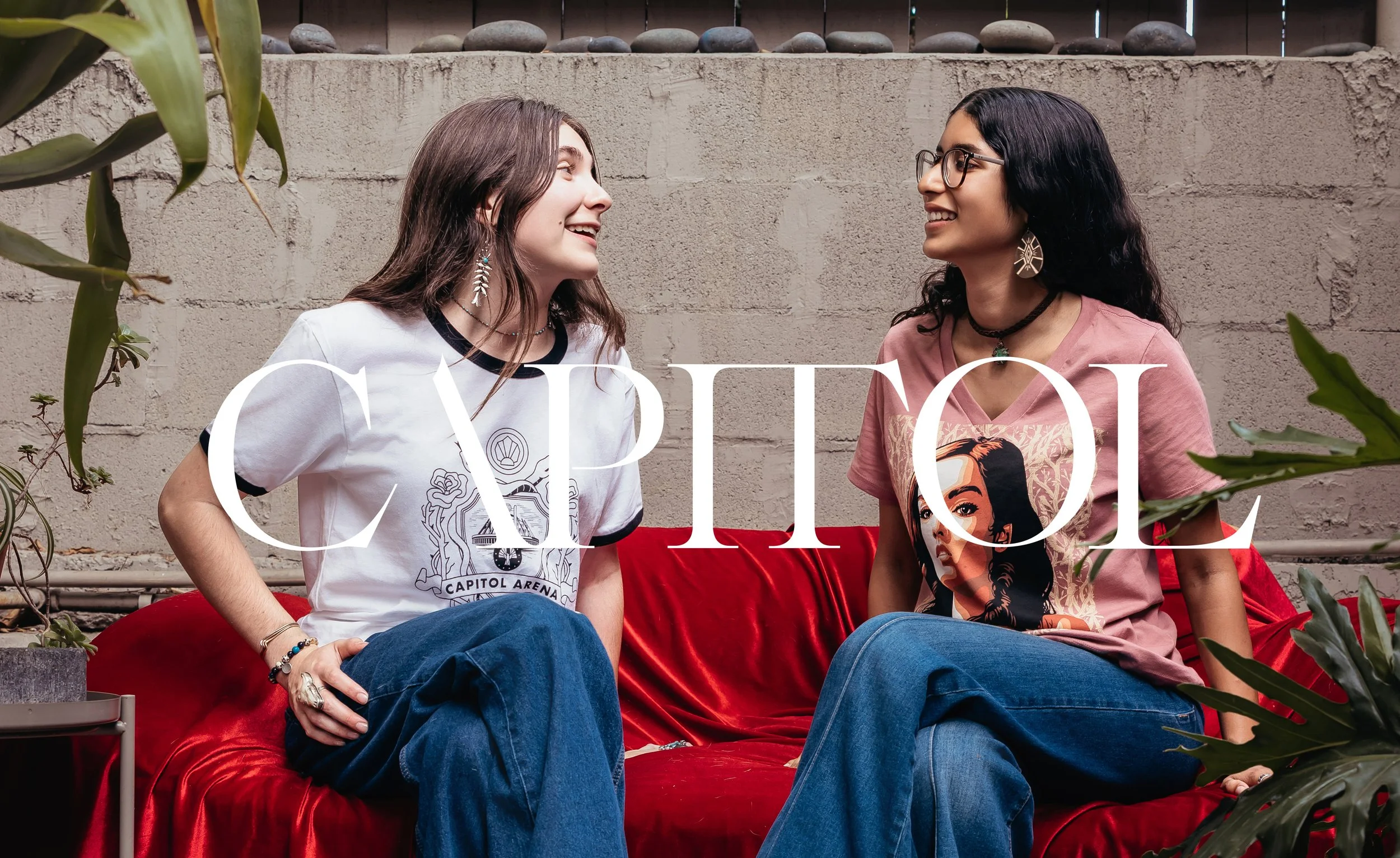 Two women sitting on a red sofa facing each other, smiling and engaged in conversation, with plants and a concrete wall in the background.