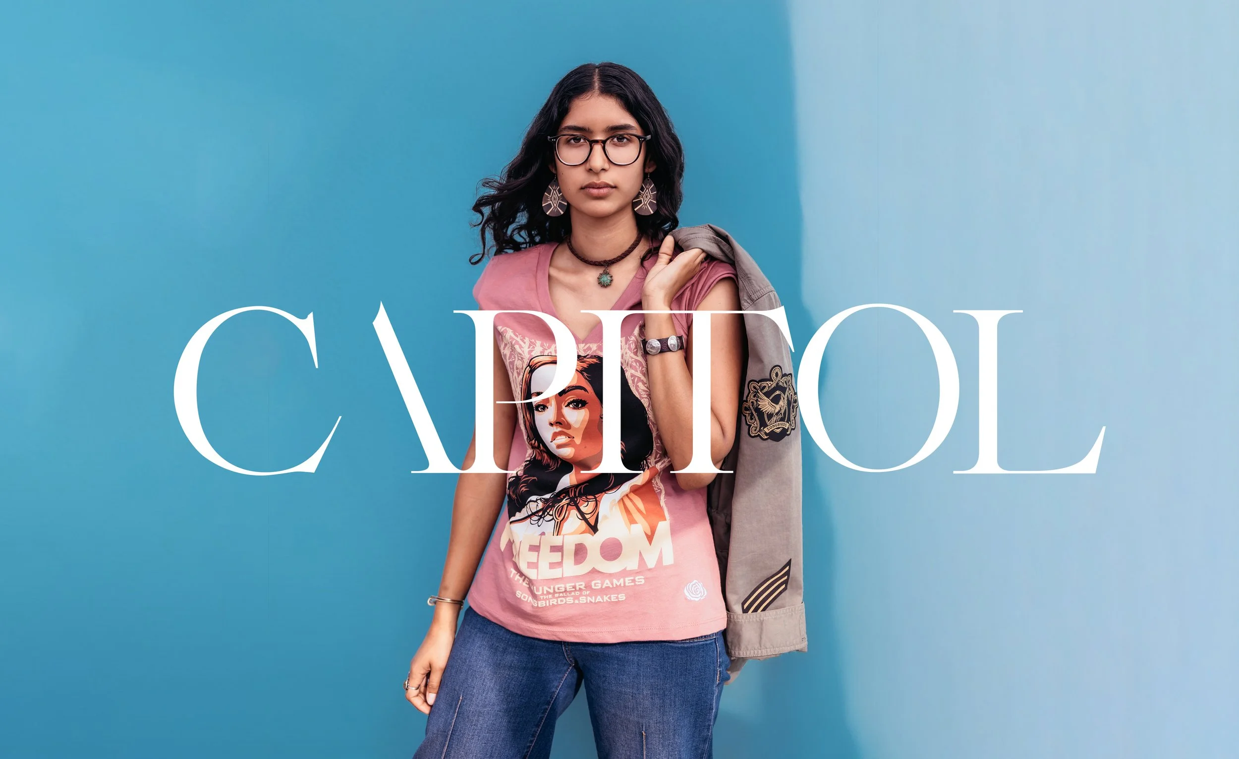 A young woman with black wavy hair, glasses, and hoop earrings poses against a two-tone blue background. She is wearing a pink graphic T-shirt with a woman’s face and the words "FREEDOM" and "THE YOUNGER GAMES," dark jeans, and multiple accessories, including a necklace, bracelet, and rings. She is holding a jacket over her shoulder.