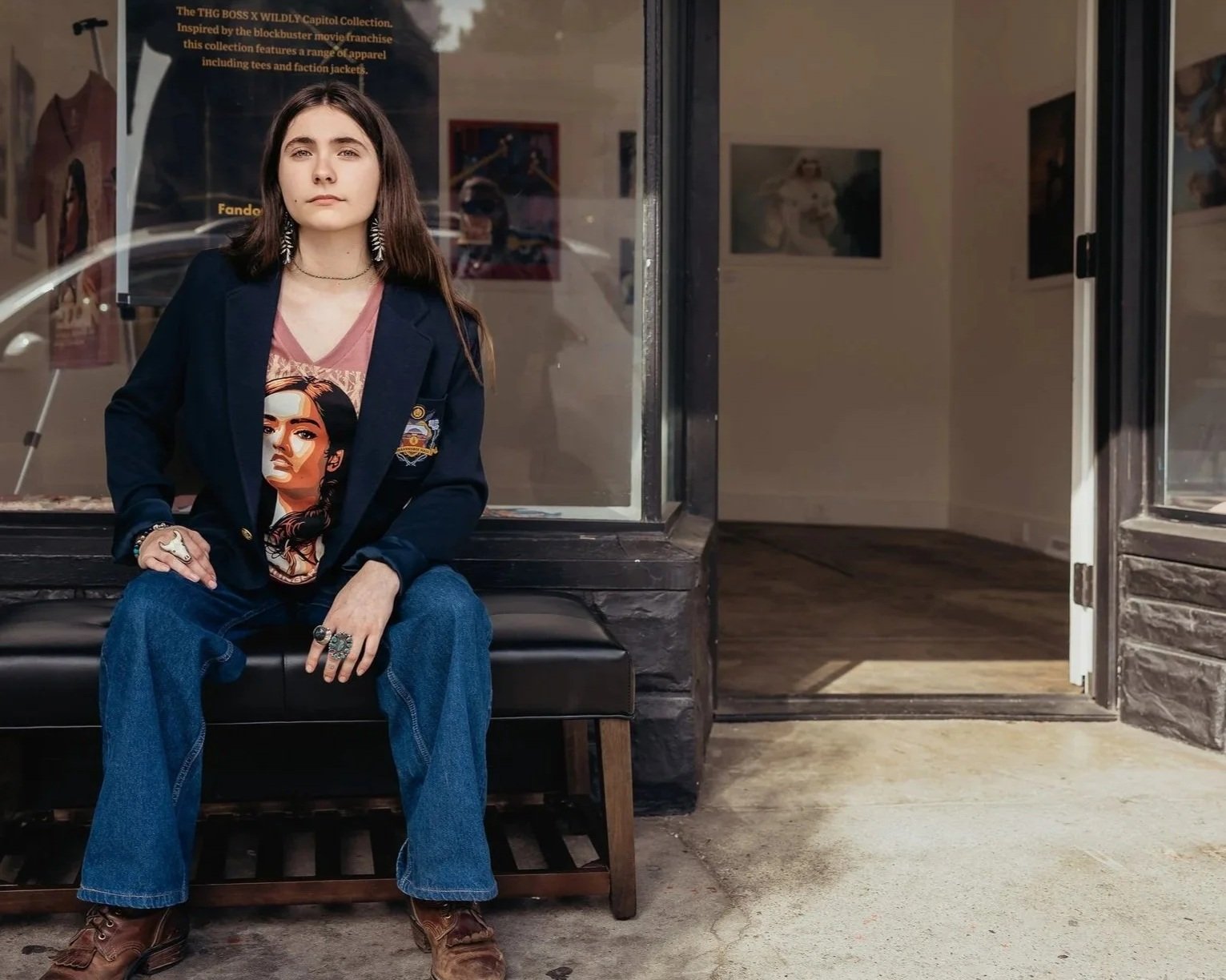 A young woman sitting on a bench outside an art gallery, wearing a dark blazer, graphic t-shirt, jeans, and boots, with jewelry, and looking directly at the camera.