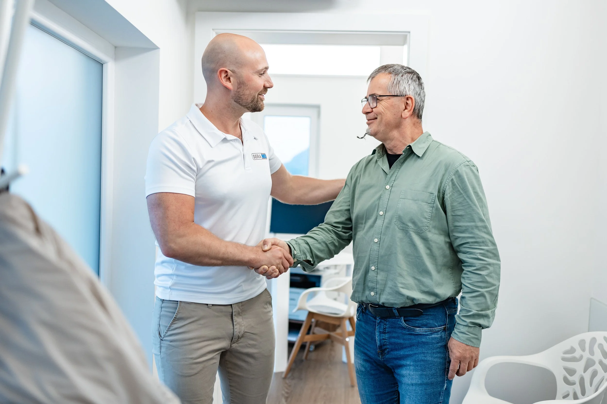 Zwei Männer schütteln sich die Hand in einem hellen Raum, einer trägt ein weißes Polo-Shirt mit Logo, der andere eine grüne Bluse, beide lächeln und schauen sich freundlich an.