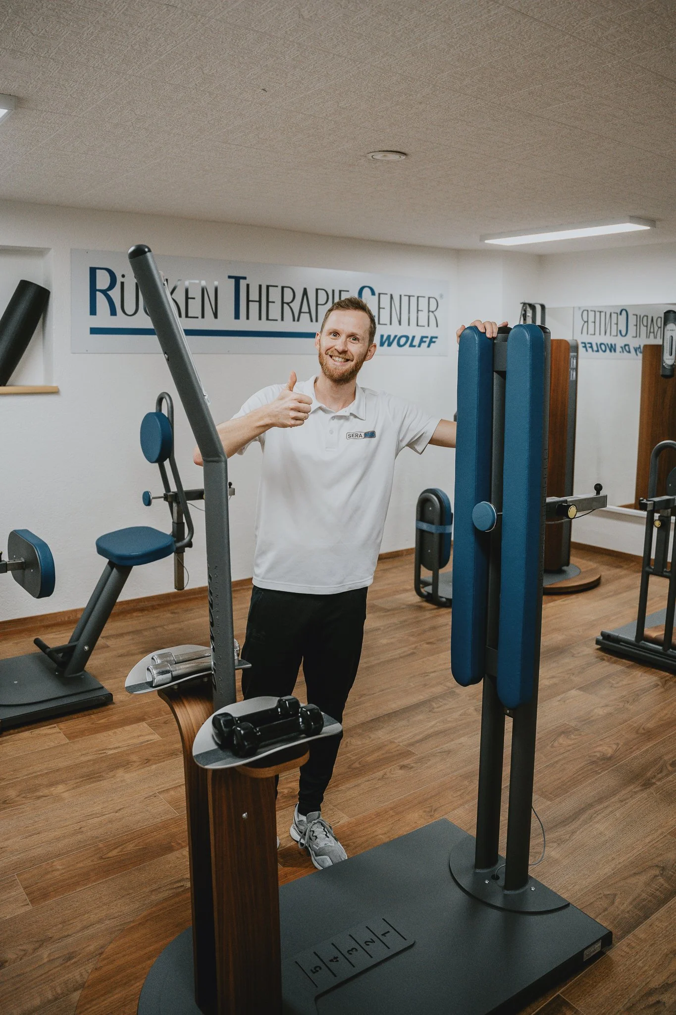 Mann in weißem T-Shirt in Fitnessstudio, der auf einem medizinischen Gerät steht, die am Boden und an der Wand stehen. Er ist amüsiert und zeigt Daumen hoch. Im Hintergrund ist ein Schild mit der Aufschrift 'Rücken Therapie Center'.