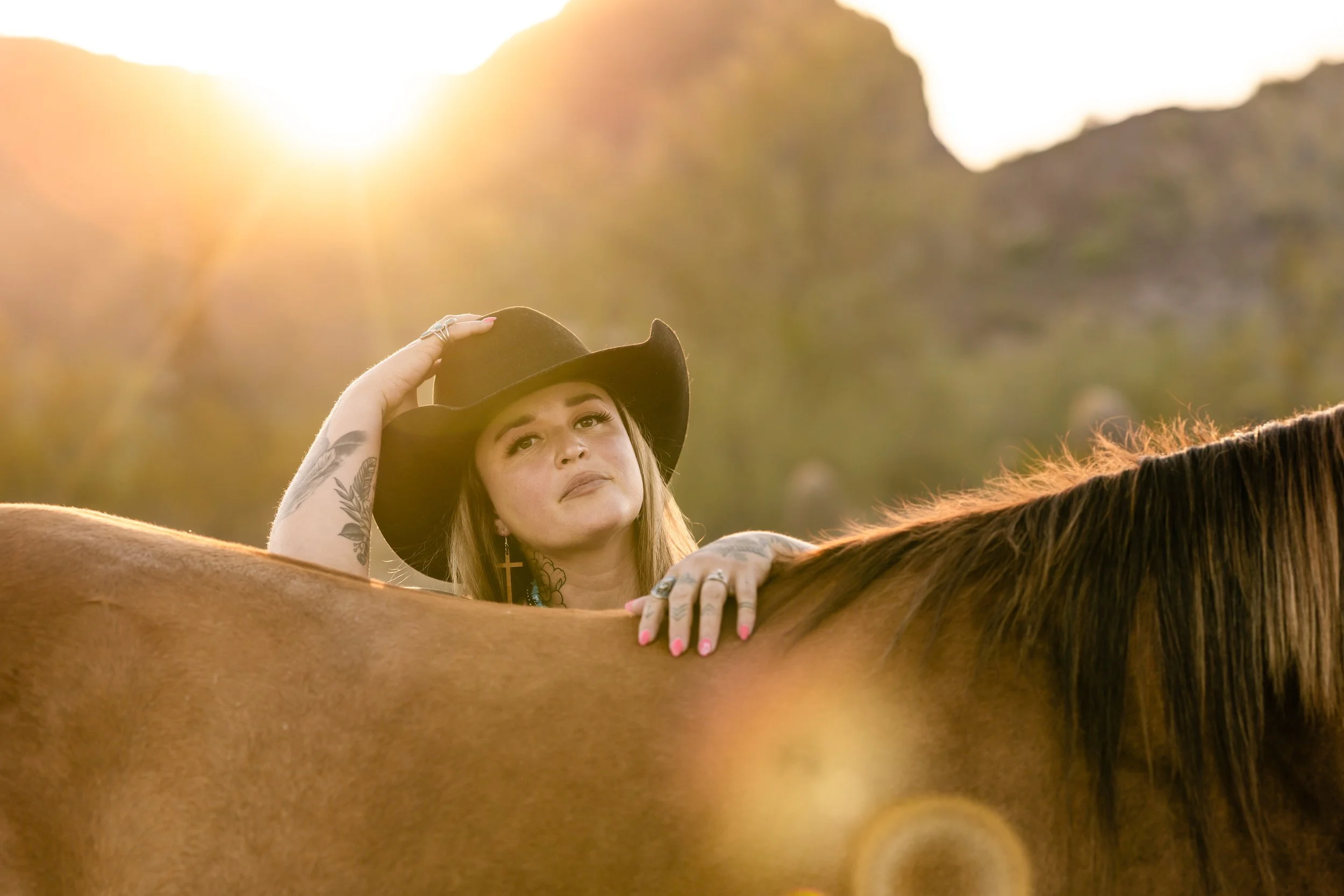 A woman with tattoos and earrings, wearing a wide-brimmed hat, standing behind a brown horse in a nature setting at sunset.