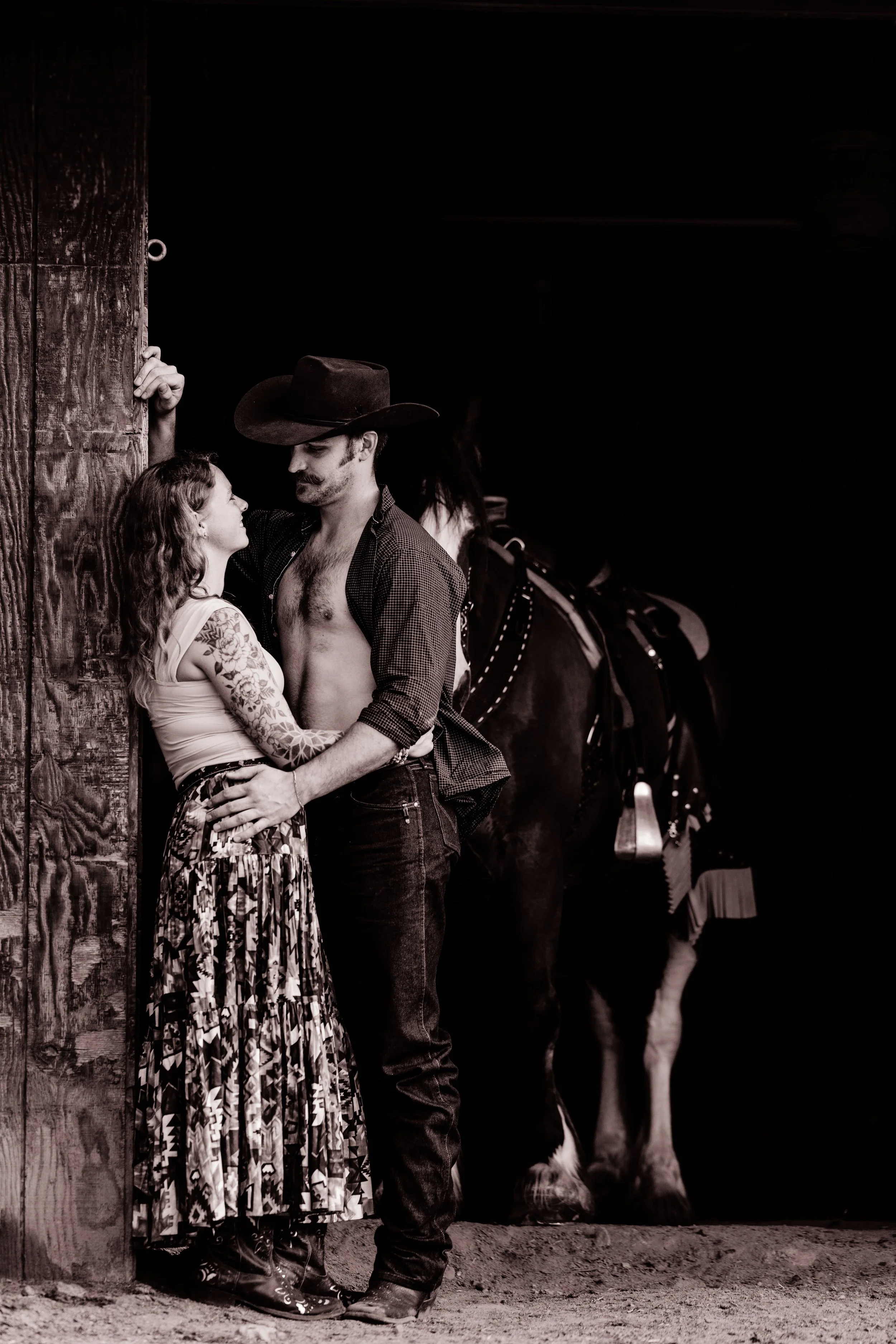 A man and woman in a romantic pose near a barn door, with a horse in the background.