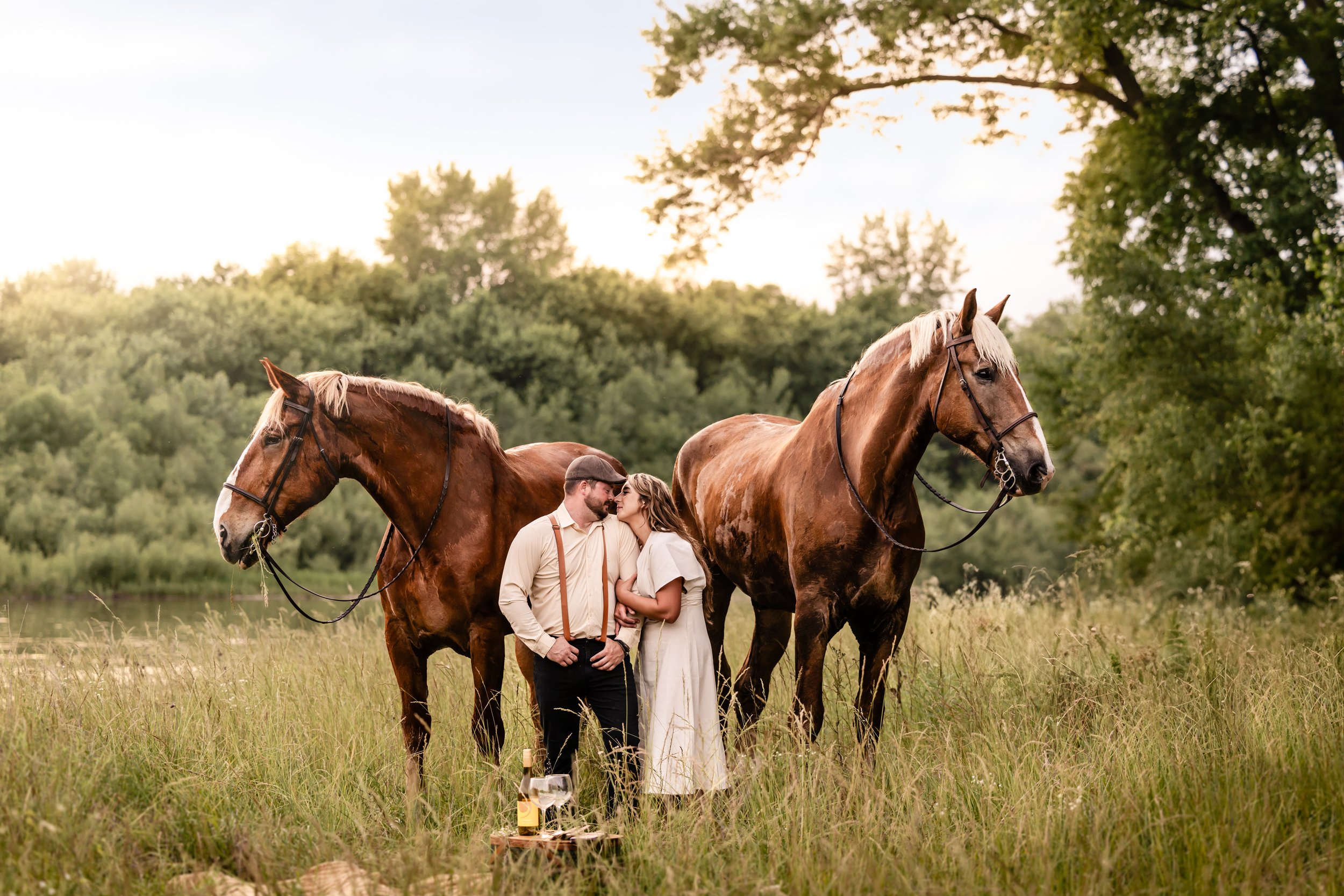 A couple standing close together in a field with two large brown horses on either side, with lush green trees and a cloudy sky in the background, during sunset.