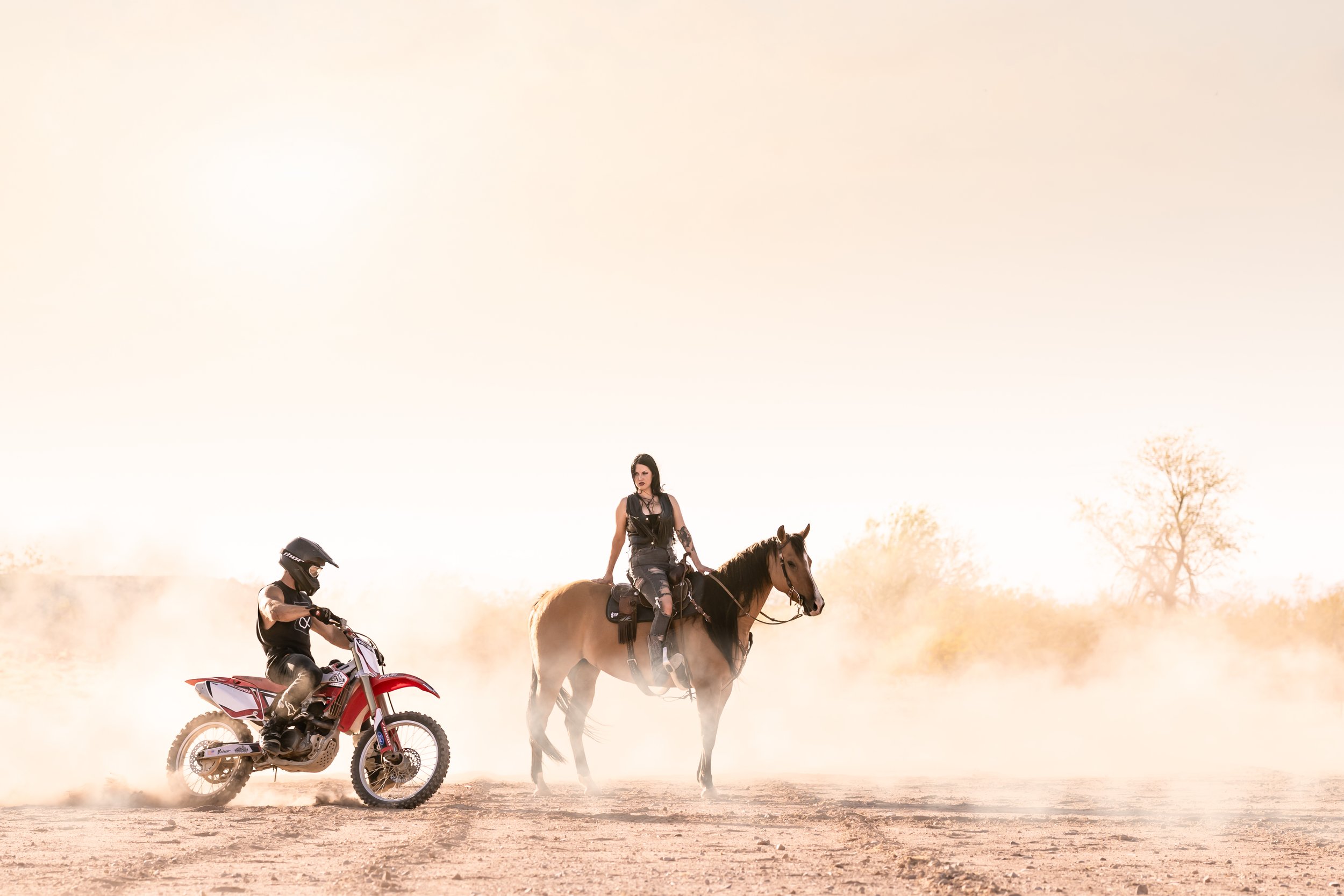 A woman riding a horse and a man on a dirt bike in a dusty, open landscape.