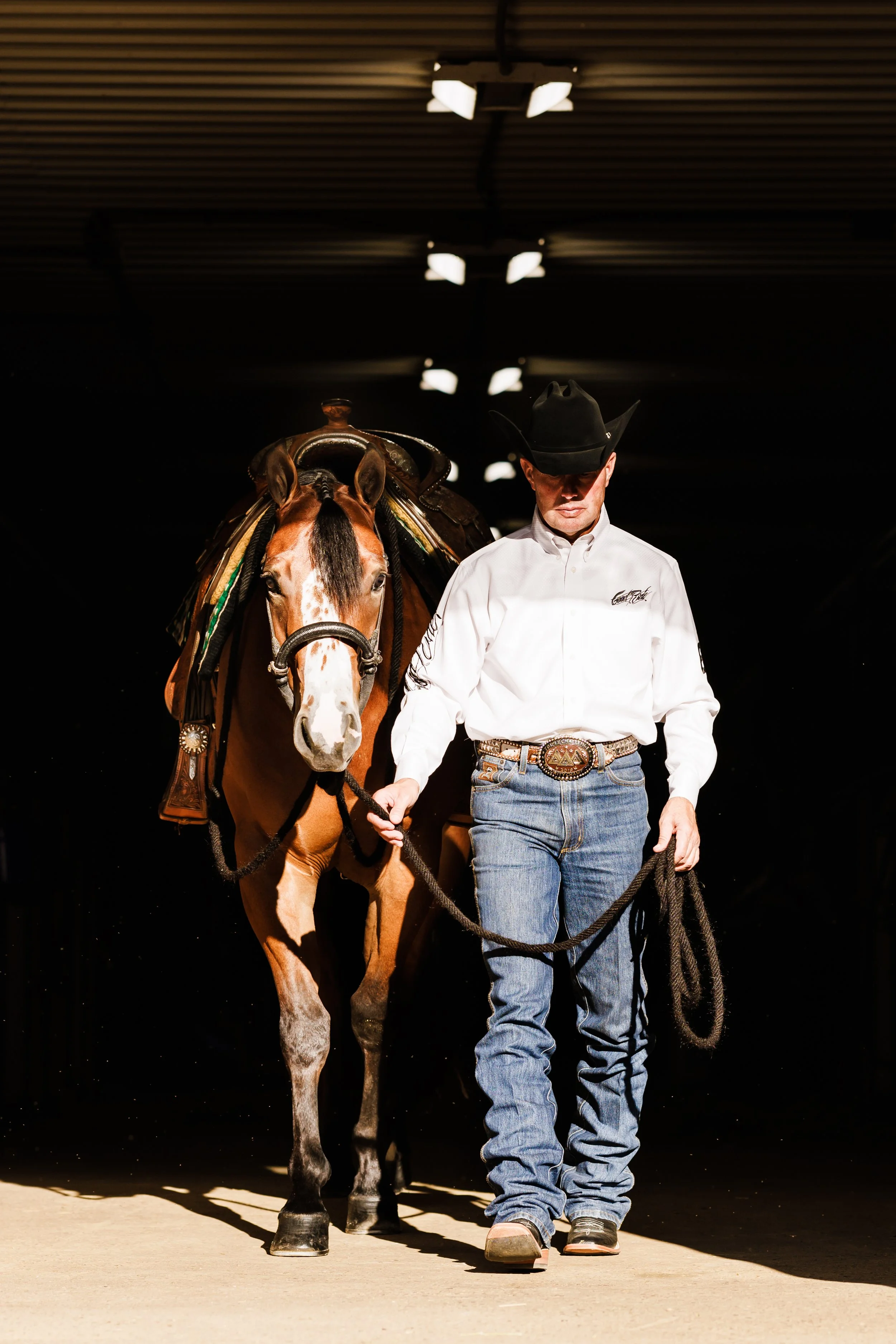 A man dressed in a white button-up shirt, blue jeans, and a cowboy hat leading a brown and white horse through an indoor facility with artificial lighting.