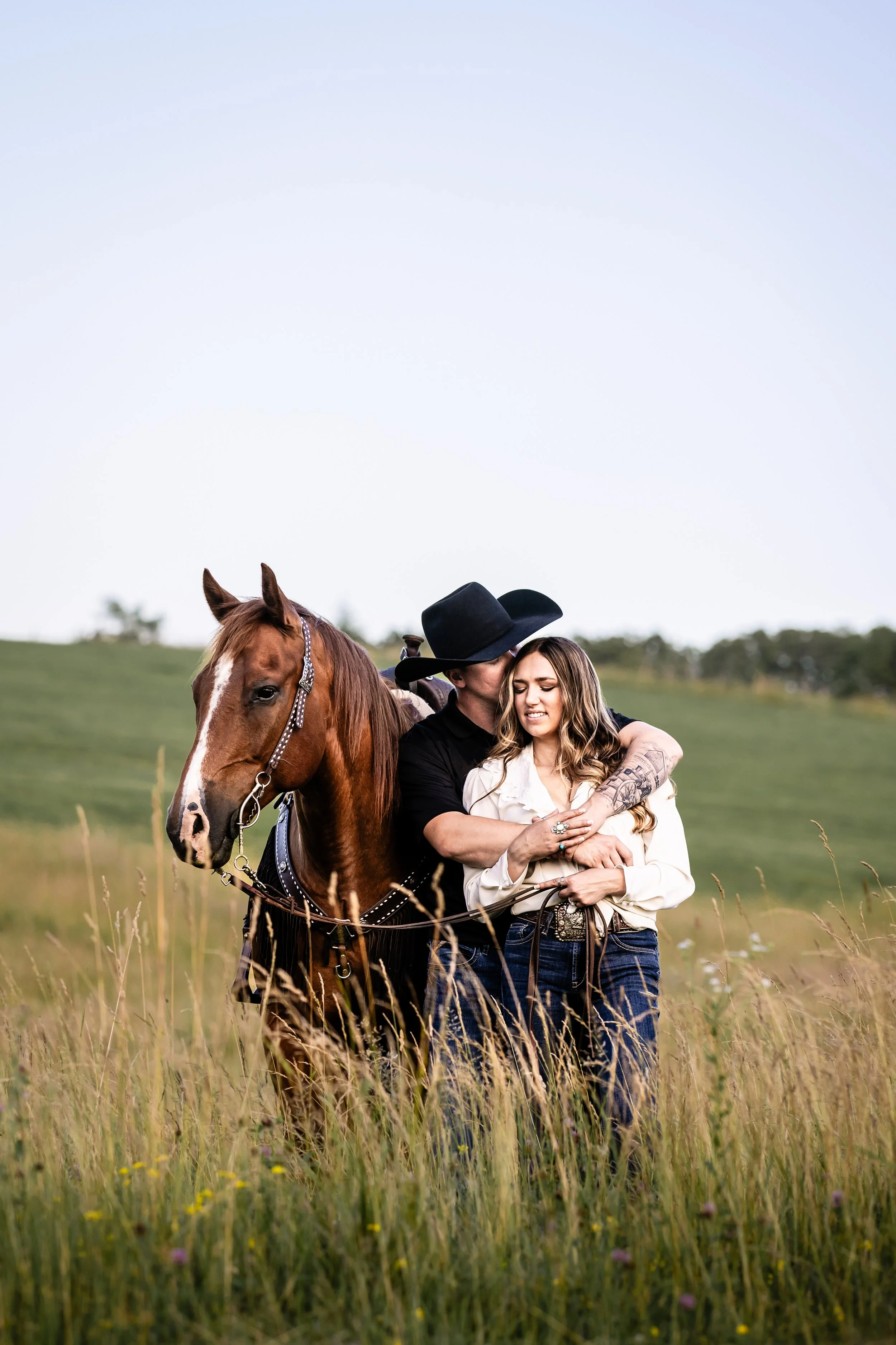 A man in a black cowboy hat and black shirt embraces a woman with long hair in a white blouse, standing next to a brown horse in a grassy field.