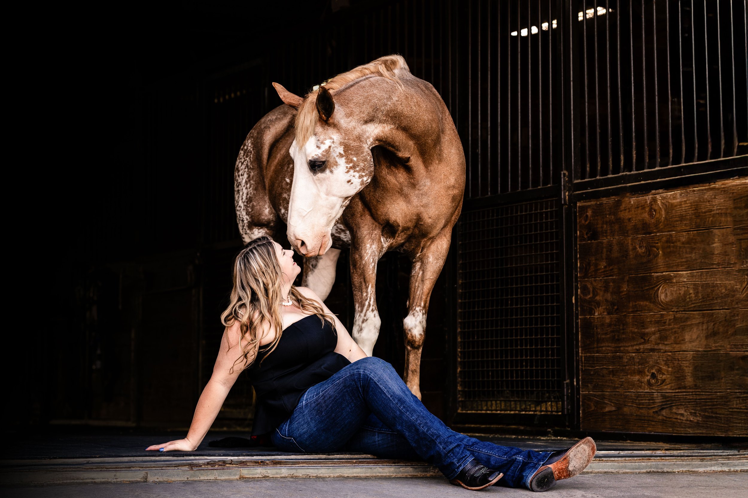 A woman with long blonde hair, wearing a black top and blue jeans, sitting on the ground inside a horse stable, looking at a large brown and white horse that is standing close to her.