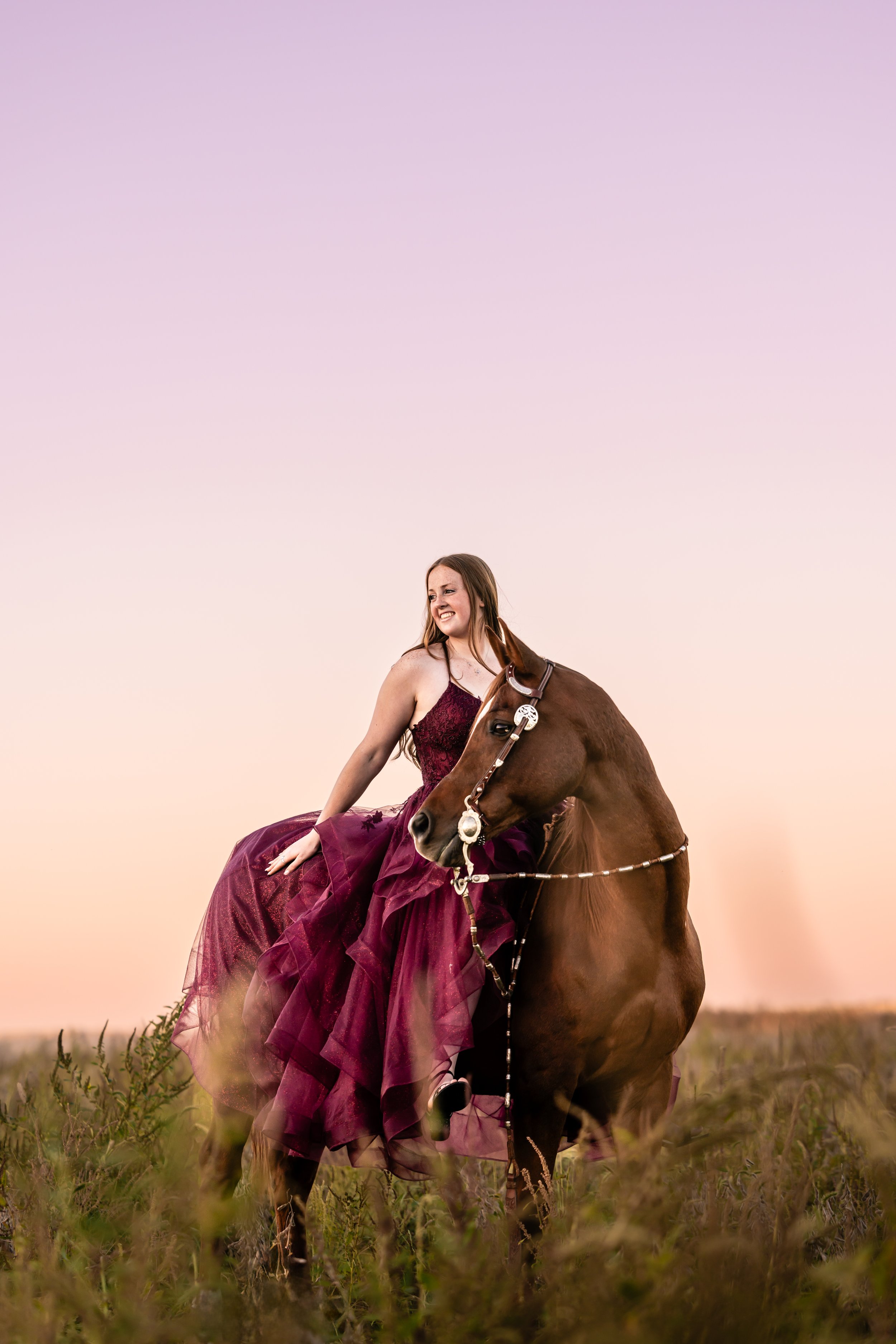 A woman in a maroon evening gown sitting on a brown horse in a grassy field during sunset.