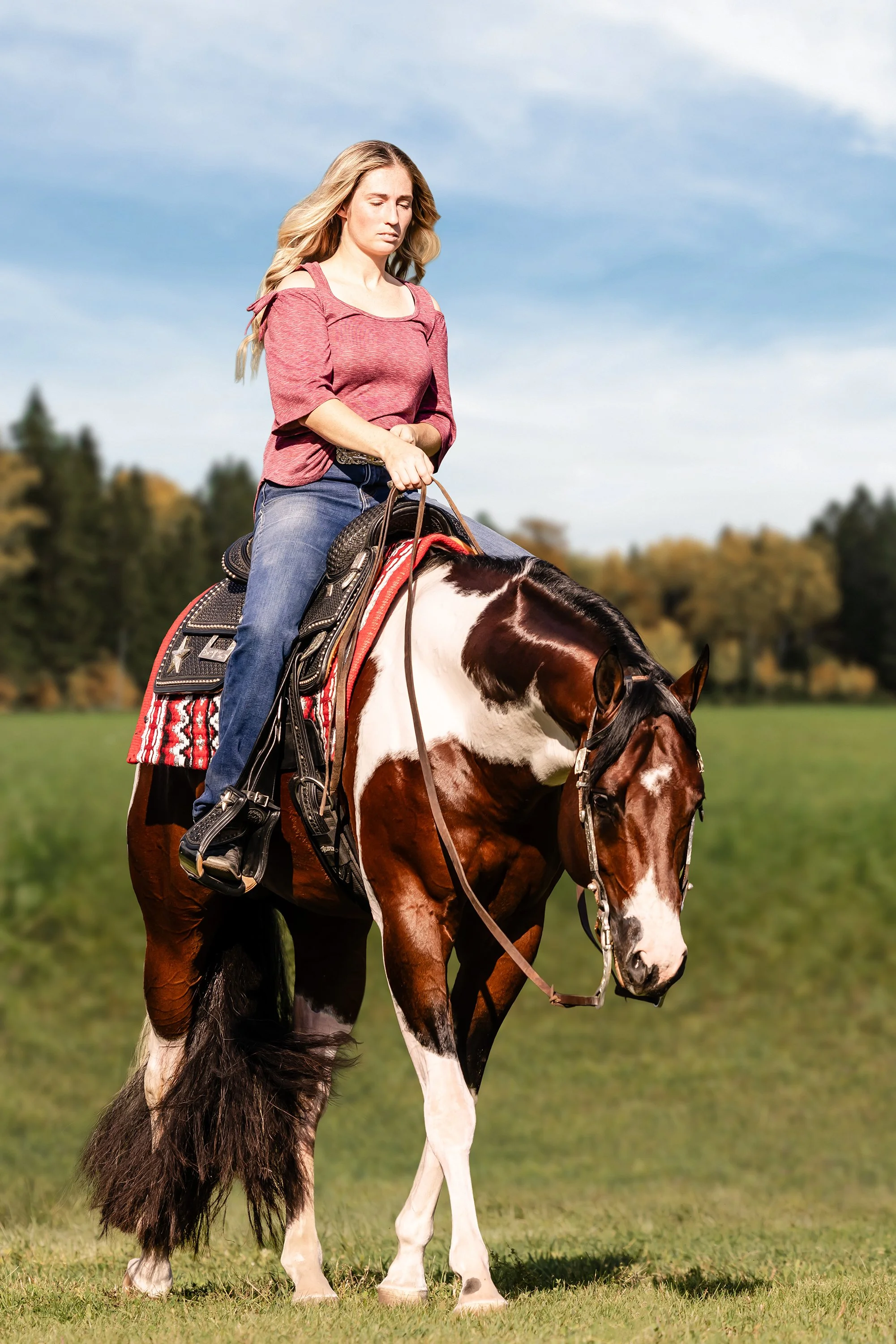 A woman riding a brown and white pinto horse in a grassy field with trees in the background.