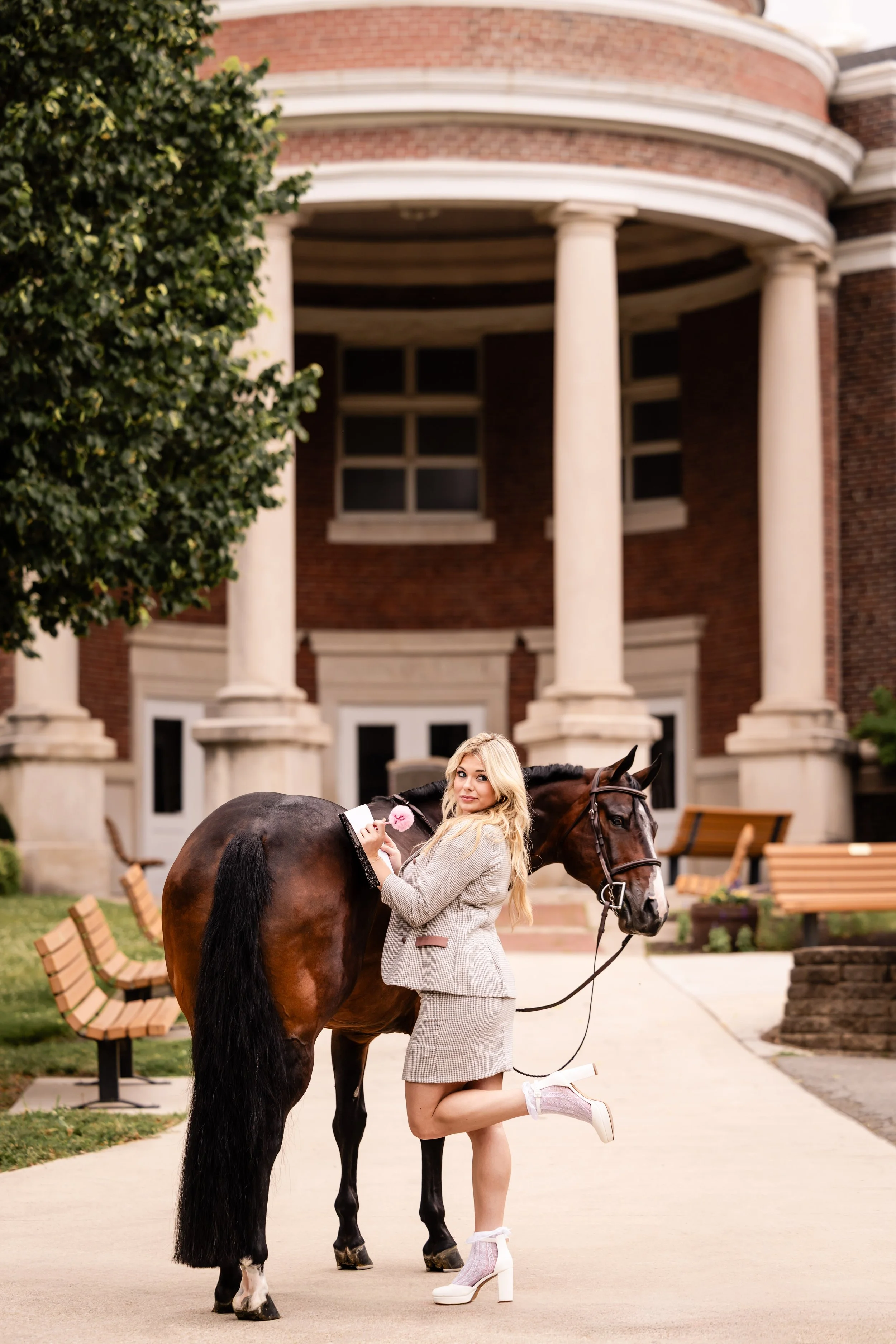 A woman in a gray suit with a skirt and white high heels stands next to a brown horse with a black mane and tail, holding a pink item, in front of a building with tall white columns and a brick facade.