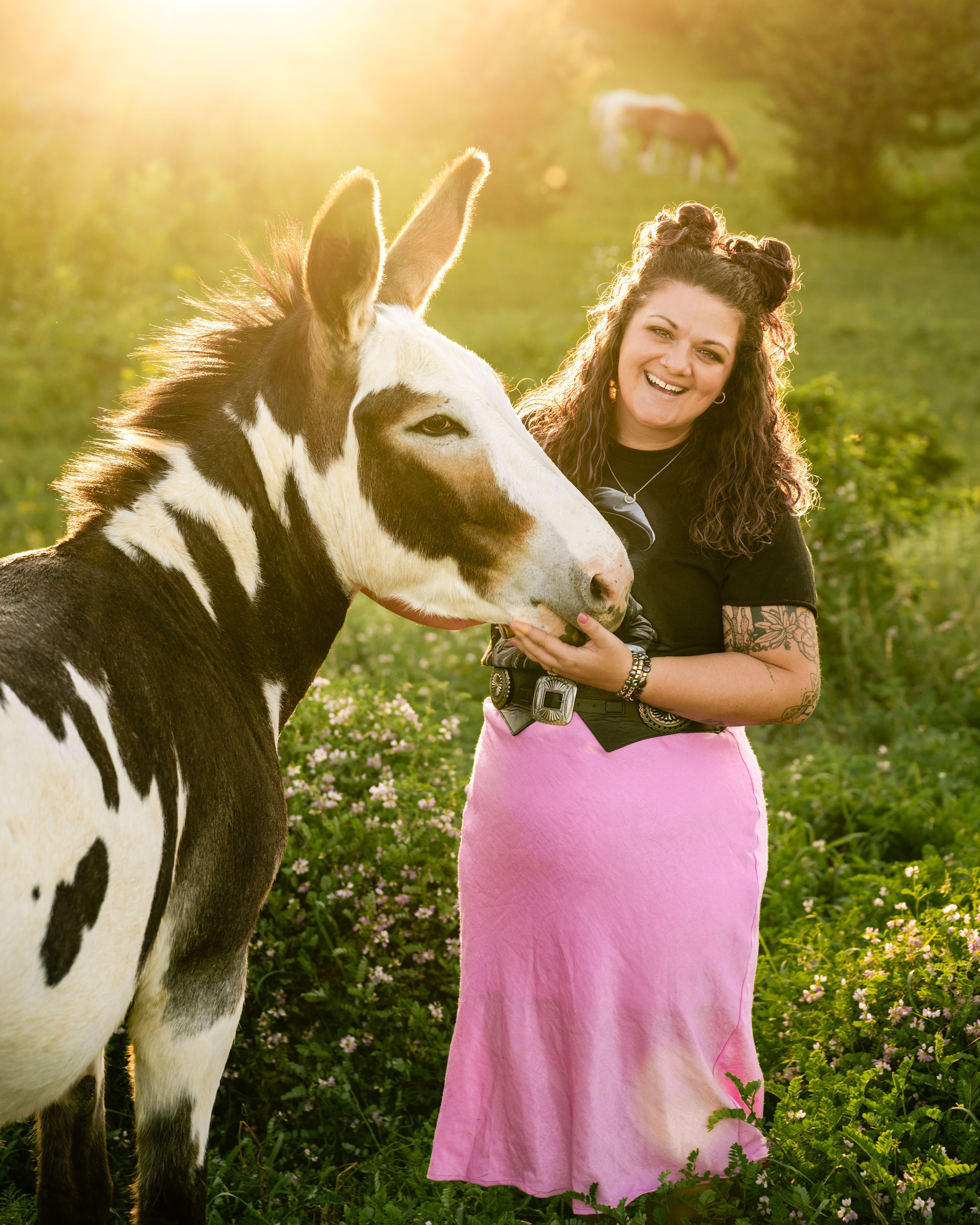 A woman with curly hair smiling and holding a black and white horse in a green field with small purple flowers, during golden hour with two other horses grazing in the background.