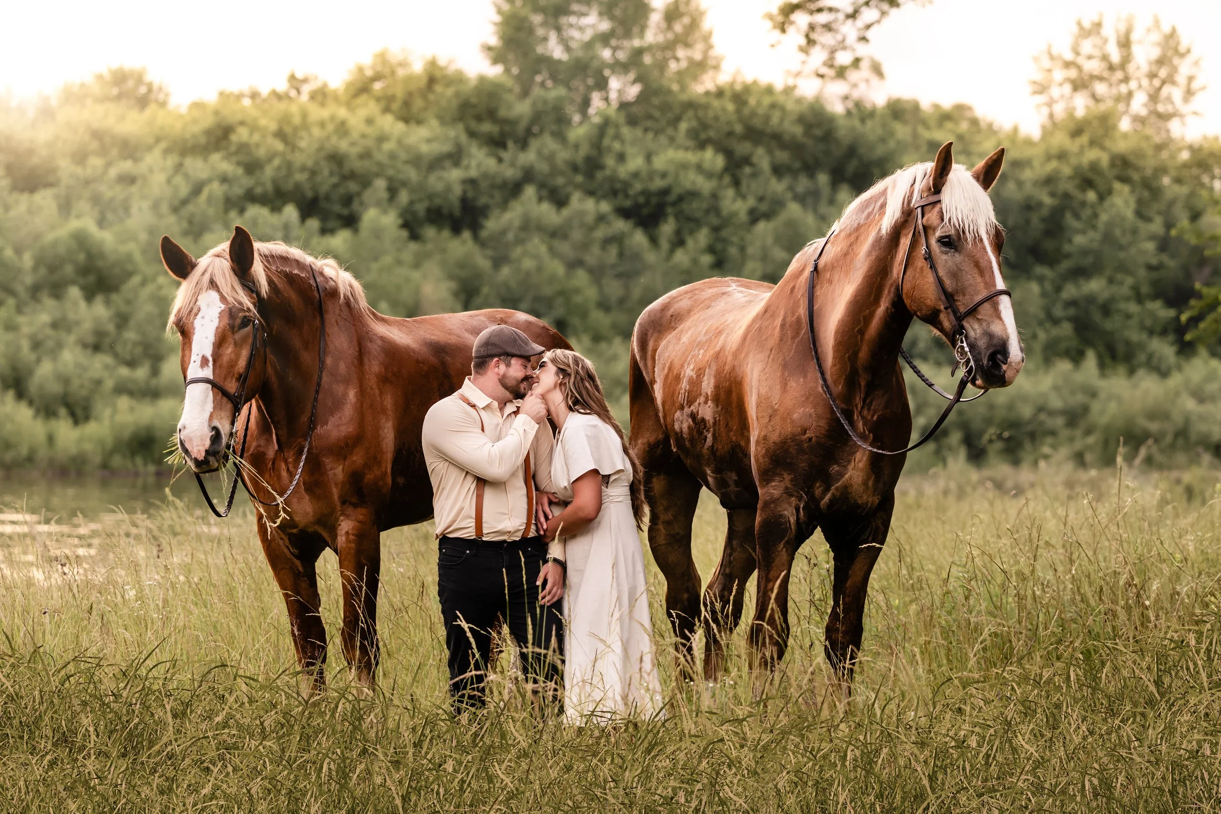 A couple stands in a grassy field between two large brown horses, sharing an intimate moment with their noses touching. The background features lush green trees under a setting sun.