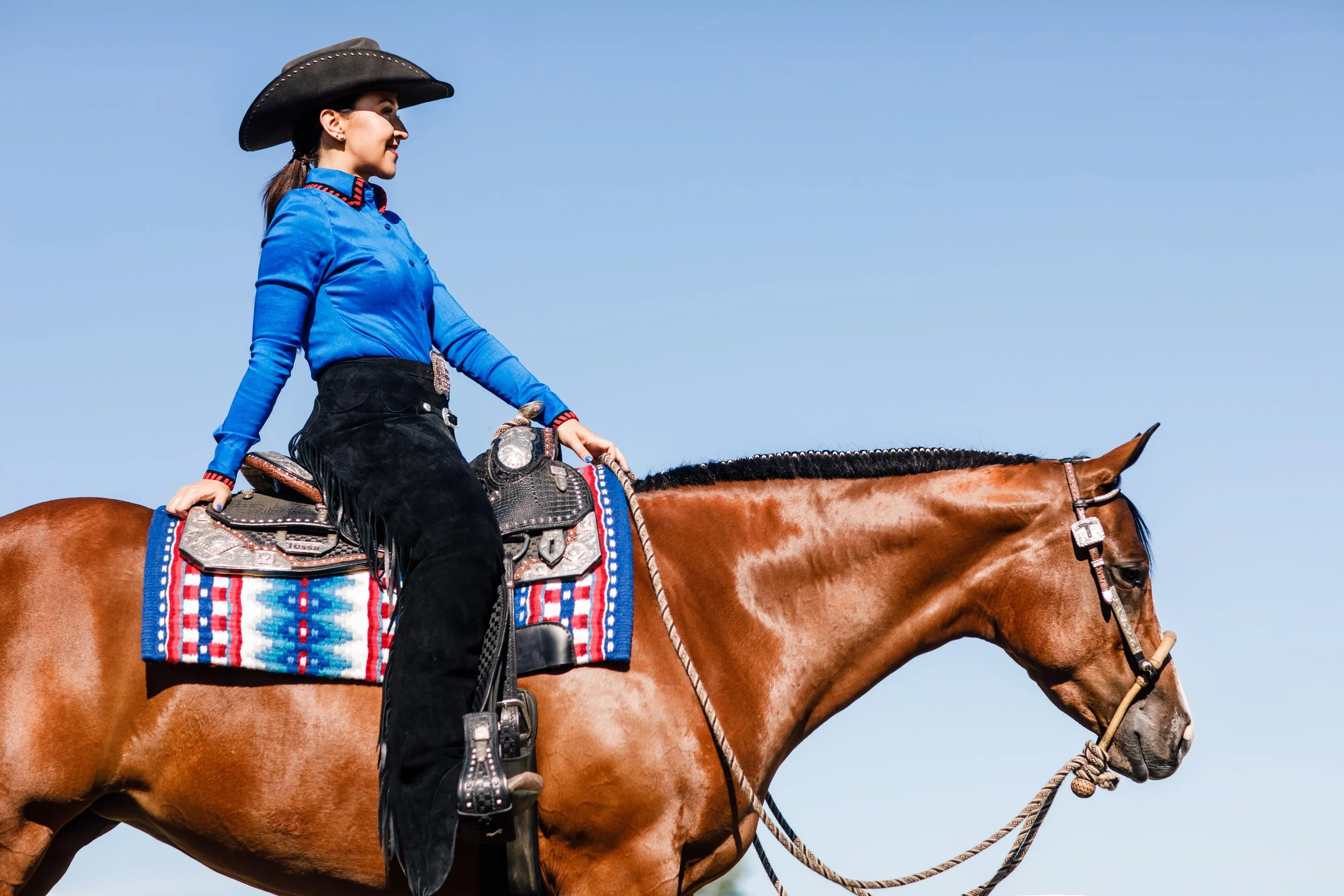 Woman riding a brown horse with a colorful saddle, wearing a blue shirt, black pants with fringe, and a cowboy hat against a clear blue sky.