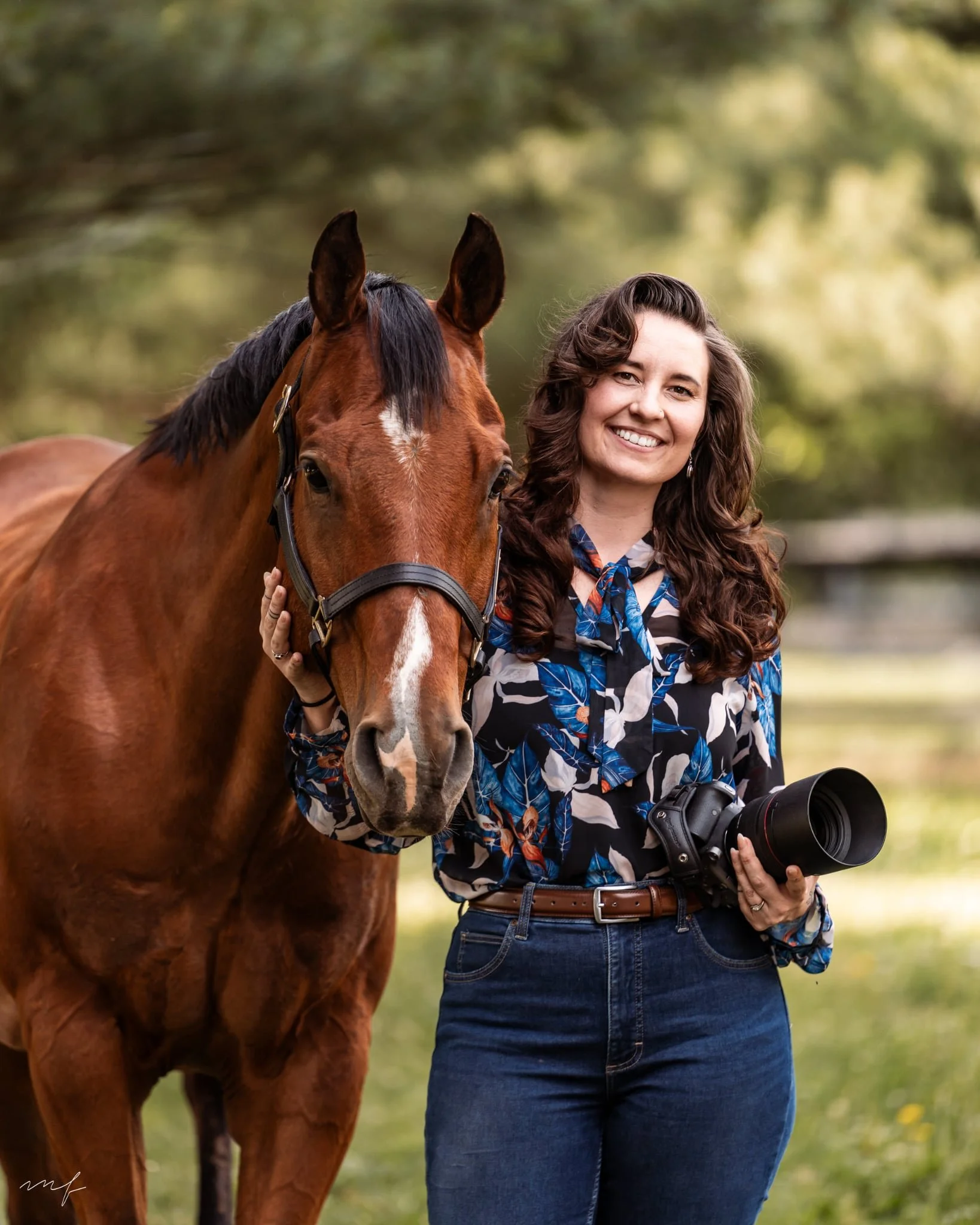 A woman with long, curly brown hair smiling while standing outdoors with a brown horse. She is holding a camera in her right hand, and the horse is wearing a halter.