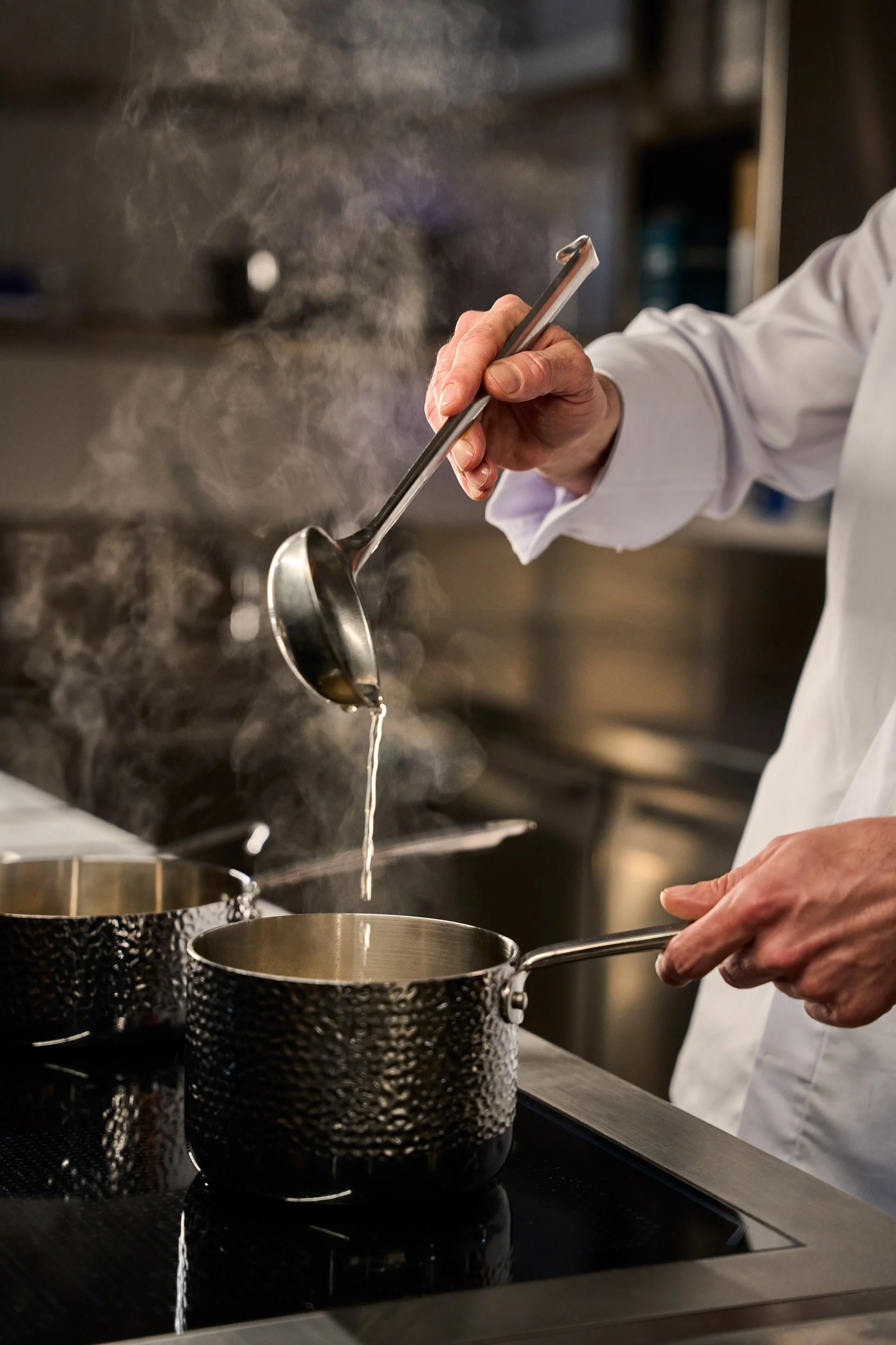 Fotografia food professionale: chef versa brodo con mestolo tra vapore in cucina professionale di ristorante a Milano — Cardinali Studio