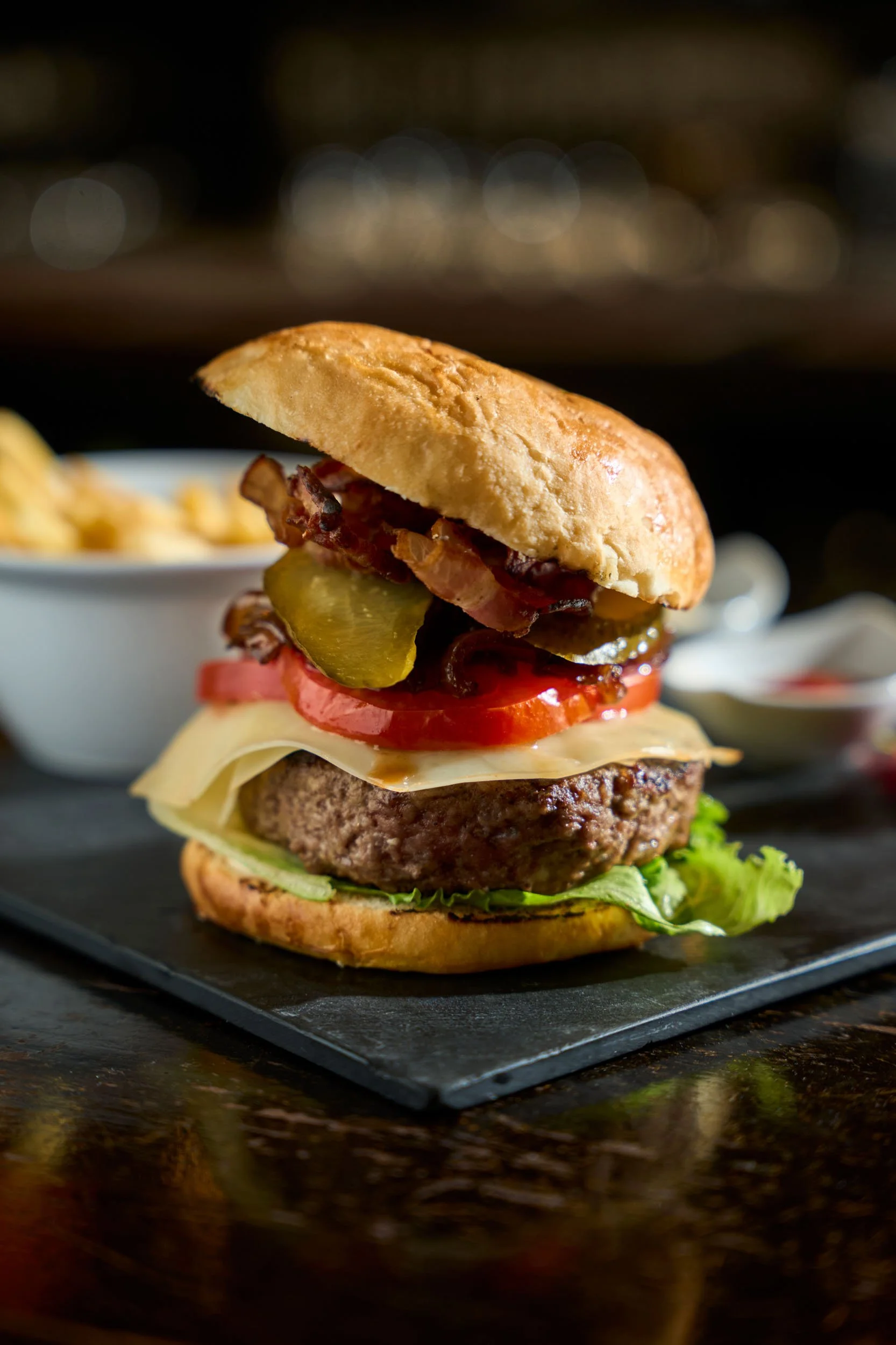 Hamburger gourmet con bacon, formaggio e patatine fritte, fotografato in primo piano su un piatto di ardesia. Servizio fotografico food per pub, hamburgerie e ristoranti a Milano, ideale per valorizzare i menù.