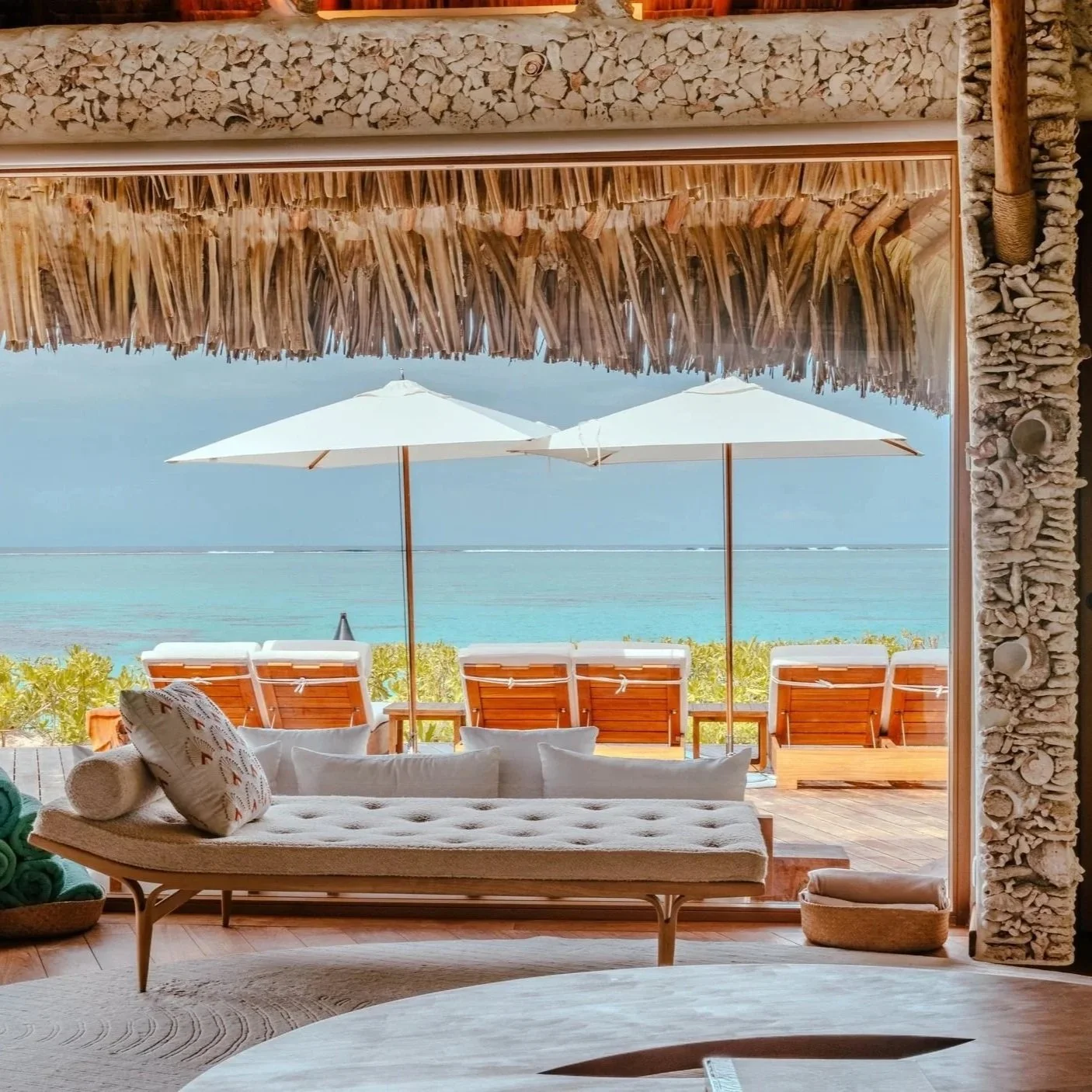 Vue depuis l'intérieur d'une villa épurée sur l'île privée préservée du Motu Nao Nao, montrant la terrasse en bois avec salon extérieur, six transats, deux parasols et une vue panoramique sur le lagon bleu turquoise.