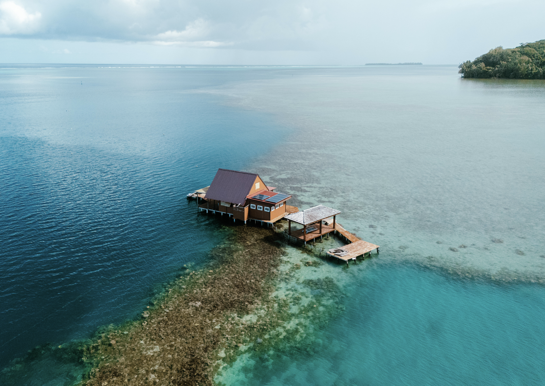 A tropical island with lush green trees and thatched-roof huts along a sandy beach. The turquoise sea has a sailboat and a person on a small sailboat with a red sail. Mountains are in the background under a partly cloudy sky.