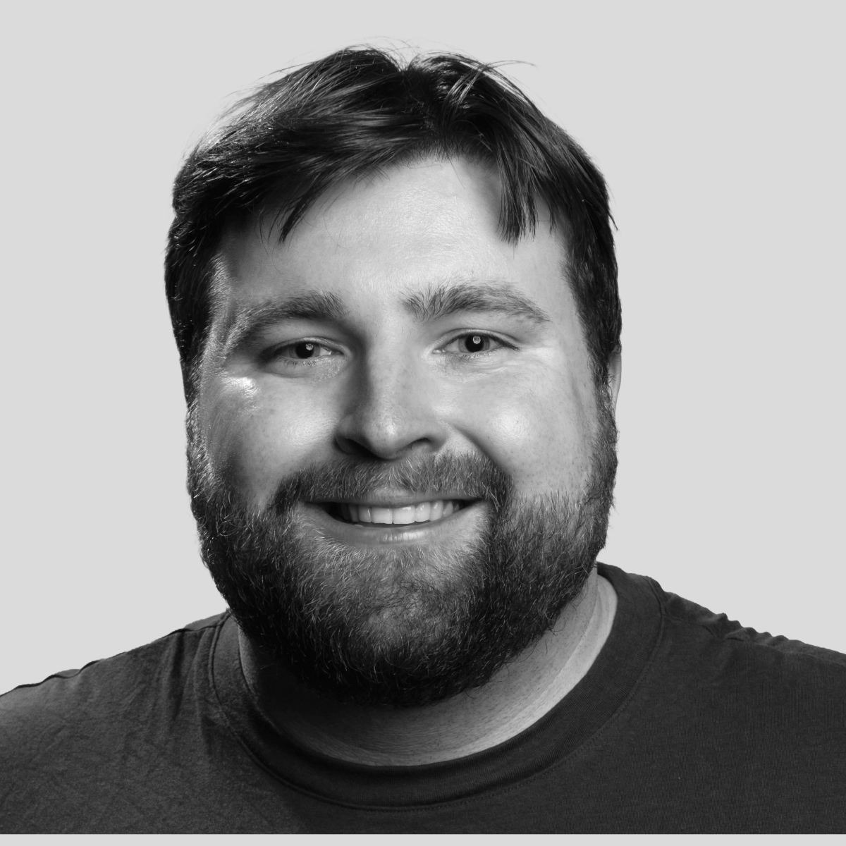 Black and white portrait of a smiling man with dark hair and a beard, wearing a dark t-shirt.