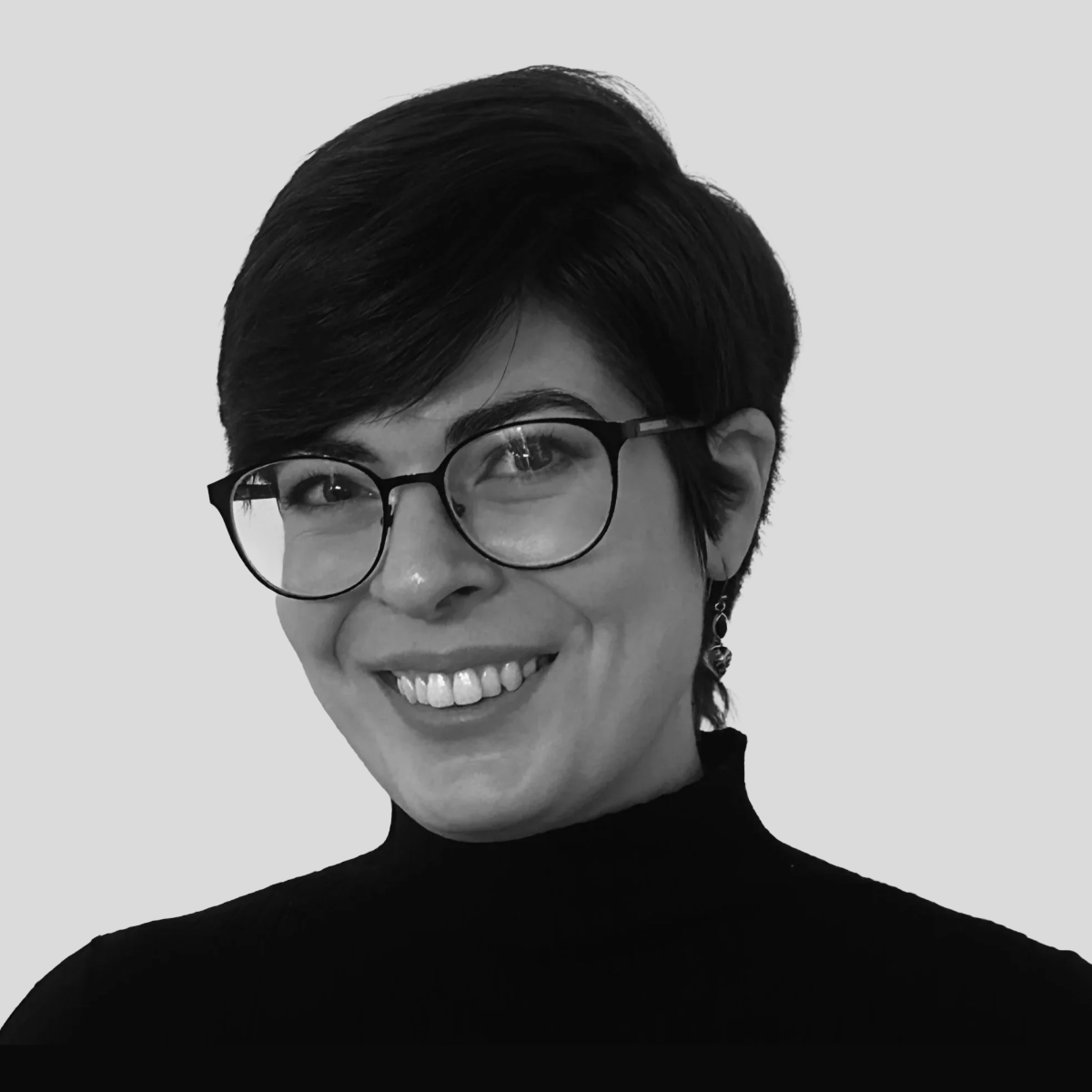 Black and white portrait of a smiling woman with short dark hair, wearing glasses, a black turtleneck, and earrings, against a plain light background.