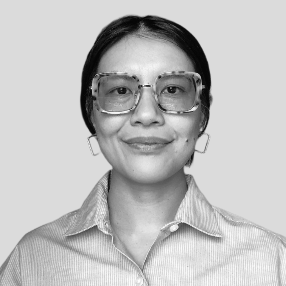A woman with short dark hair, wearing large rectangular glasses with a tortoiseshell pattern, earrings, and a striped collared shirt, smiling at the camera, black and white photo.
