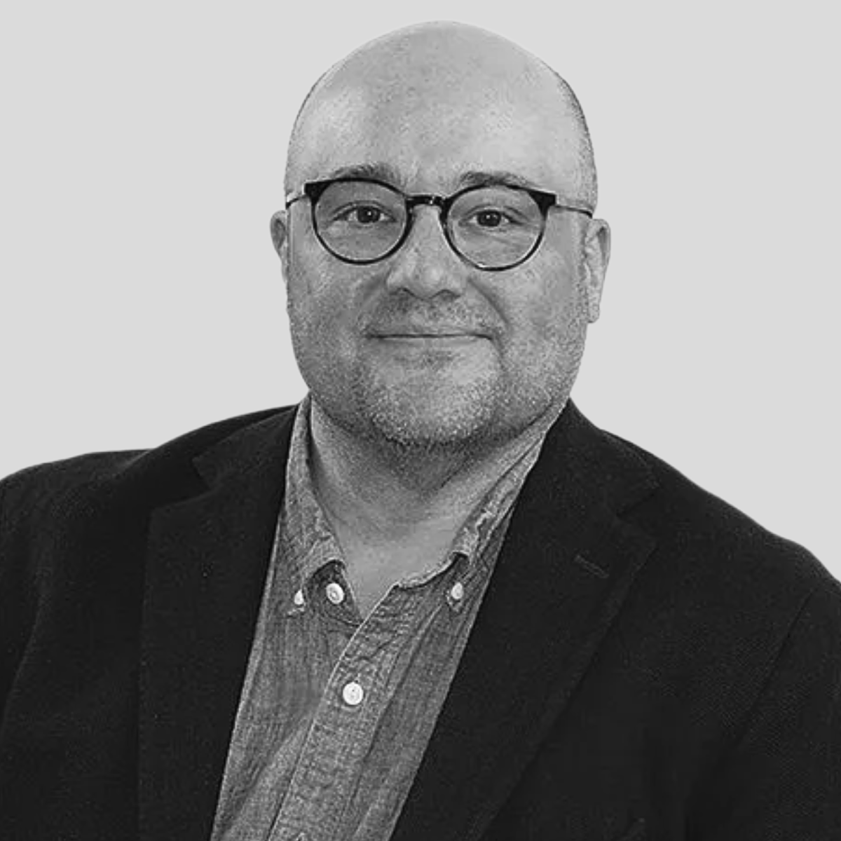 A black and white portrait photo of a man with a shaved head, glasses, a short beard, wearing a suit jacket over a collared shirt, smiling slightly, against a plain background.