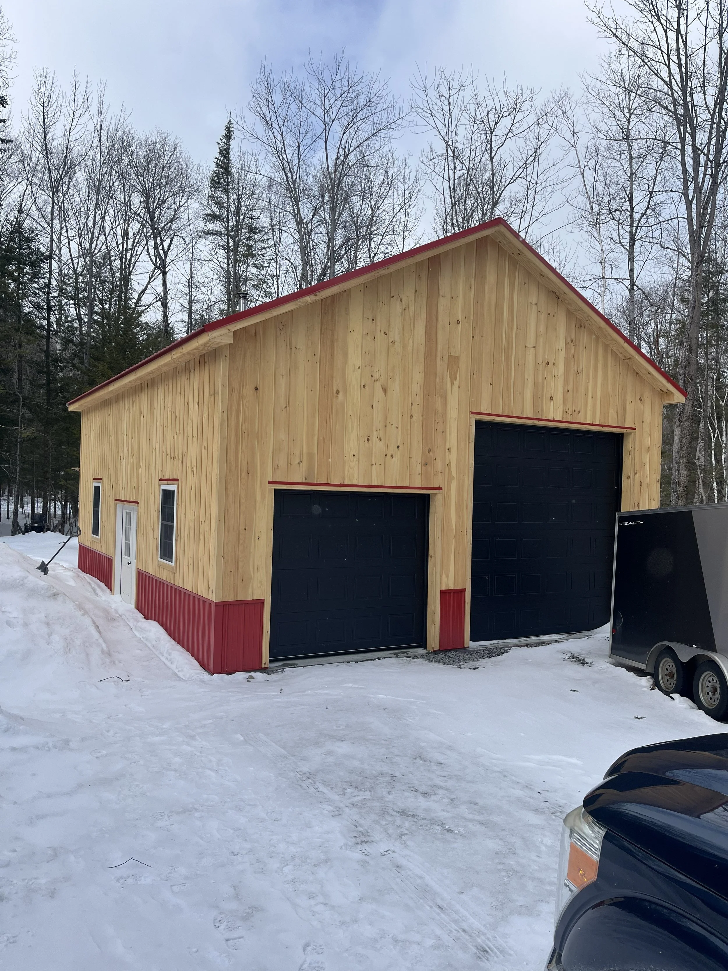 A wooden garage with black garage doors, red trim, and a snow-covered ground in a winter landscape with leafless trees in the background.